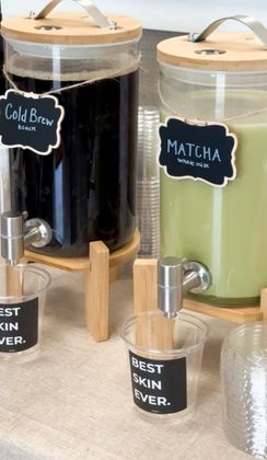Iced cold brew and matcha drinks in dispensers with wooden lids and labels, on a counter