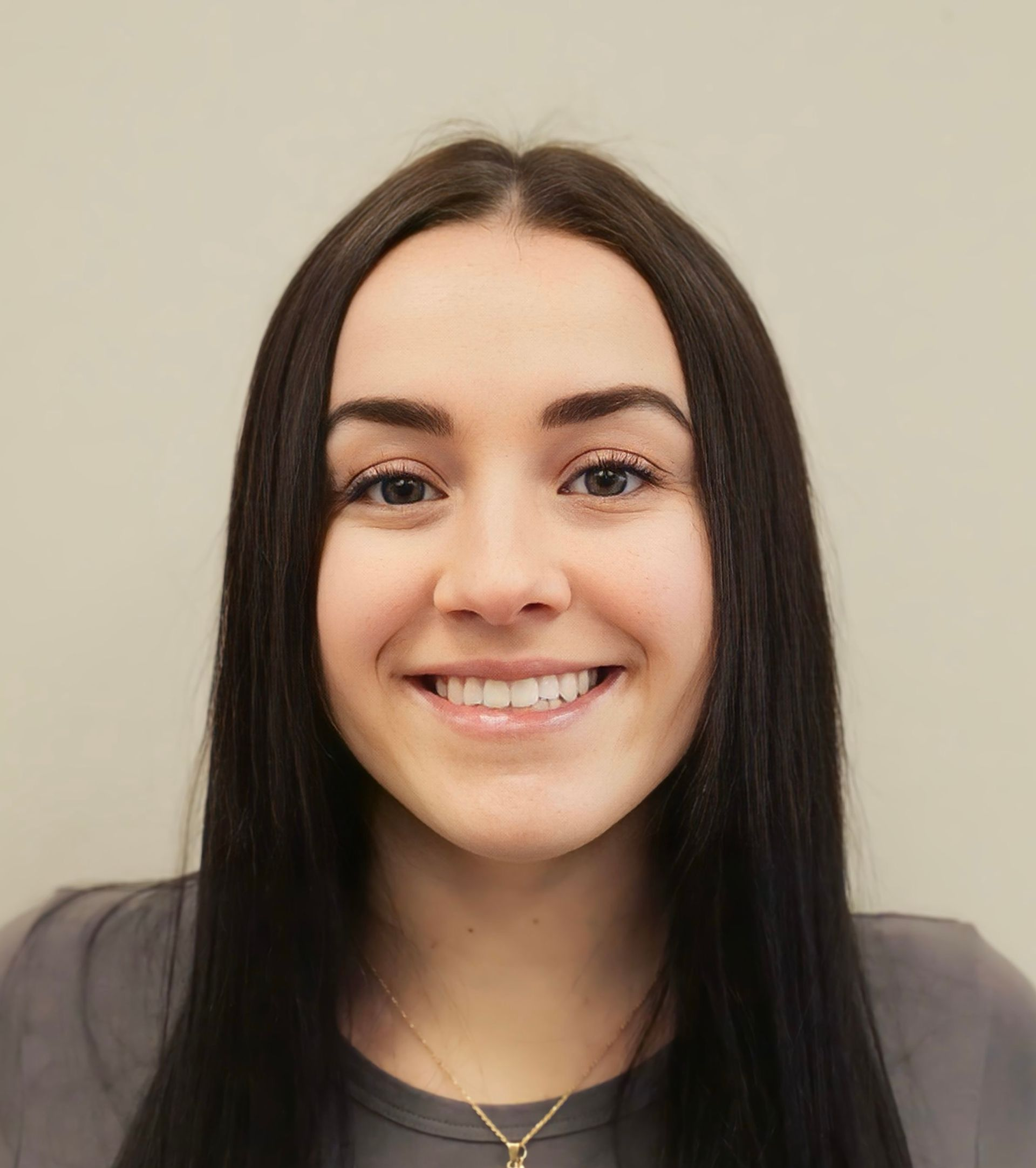 Smiling woman with long dark hair wearing a gray top and gold necklace against a beige background