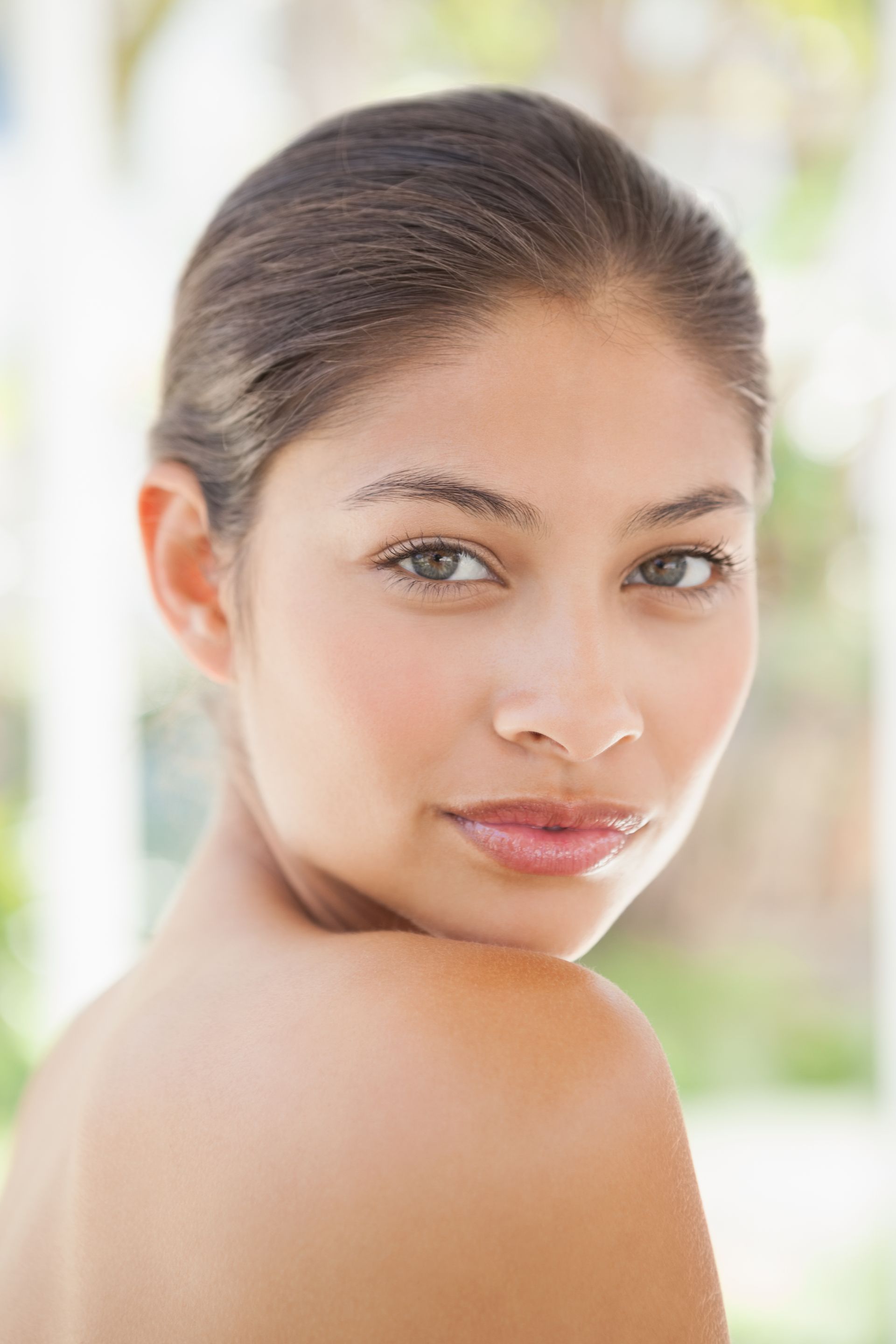 Close-up portrait of a person with tied-back hair, looking over their shoulder against a bright blurred background.
