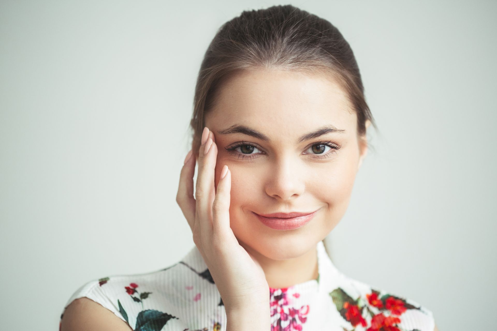 Smiling woman with hand on her cheek, wearing a white floral top against a light background