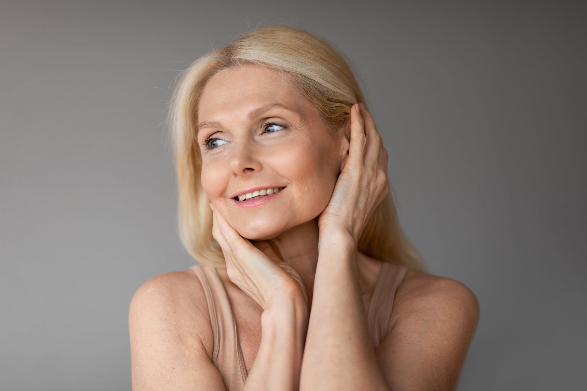 Blonde person smiling with hands on cheeks against a gray background