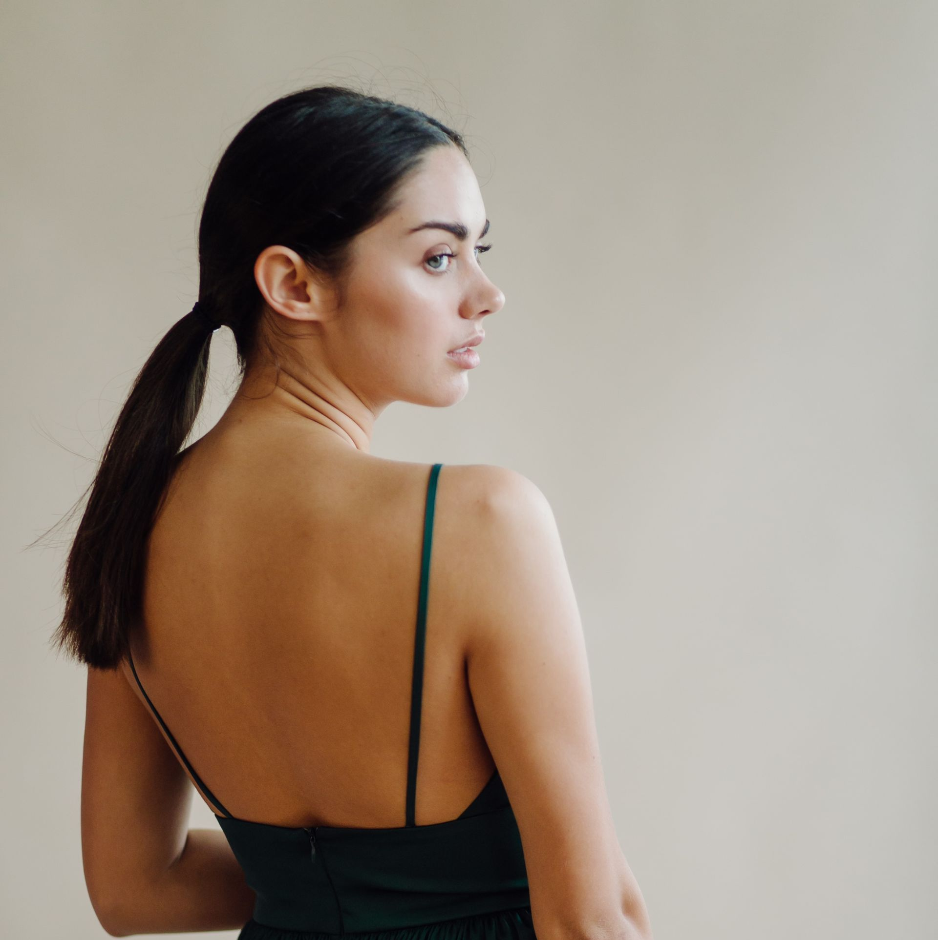 Woman in a dark green dress, seen from behind, looking over her shoulder against a neutral background