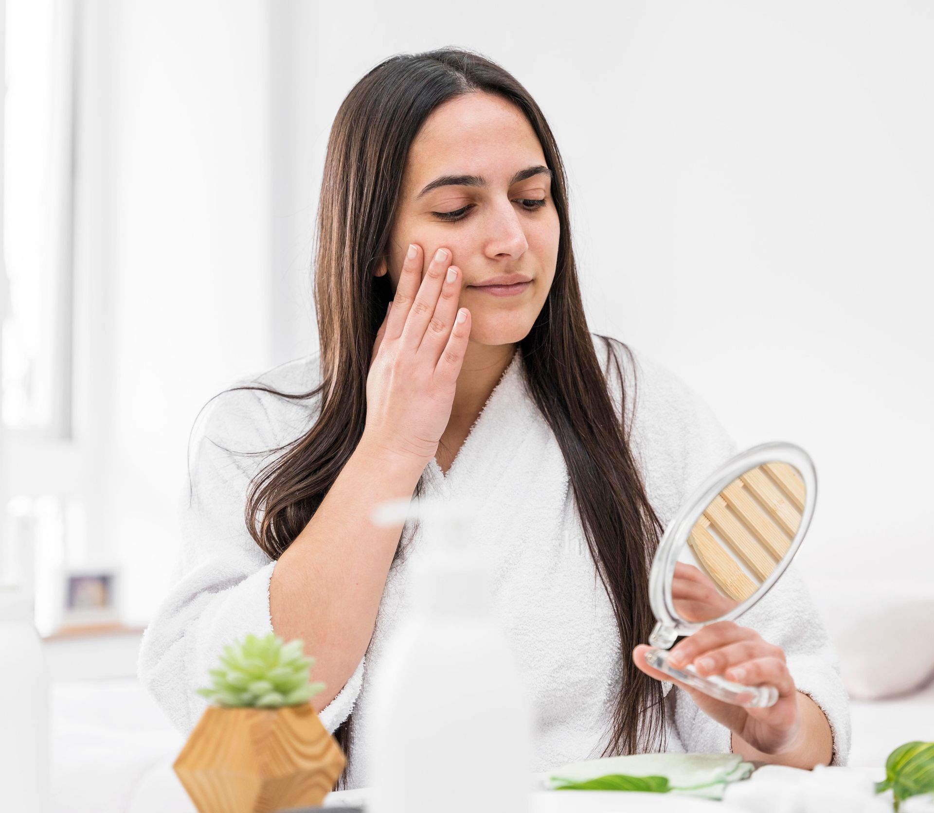 Woman applying skincare cream in a bright bathroom, holding a hand mirror and touching her cheek.