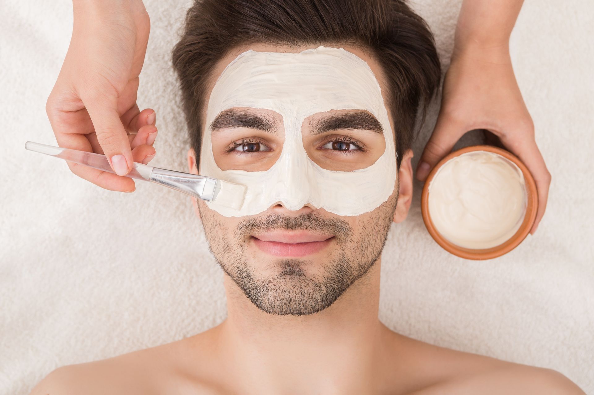 Man lying down with a white facial mask being applied using a brush, spa treatment setting