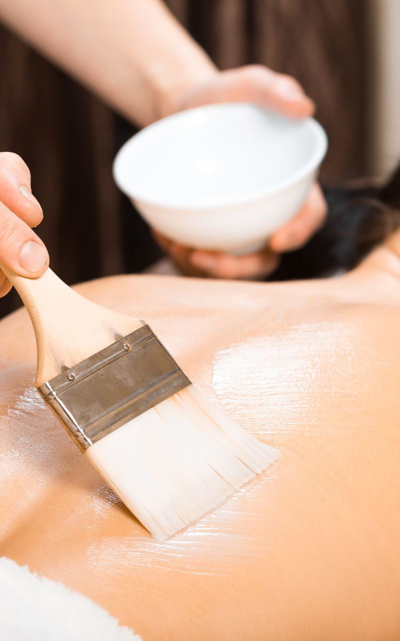 Spa therapist applying lotion to a person's back with a brush, holding a small bowl.