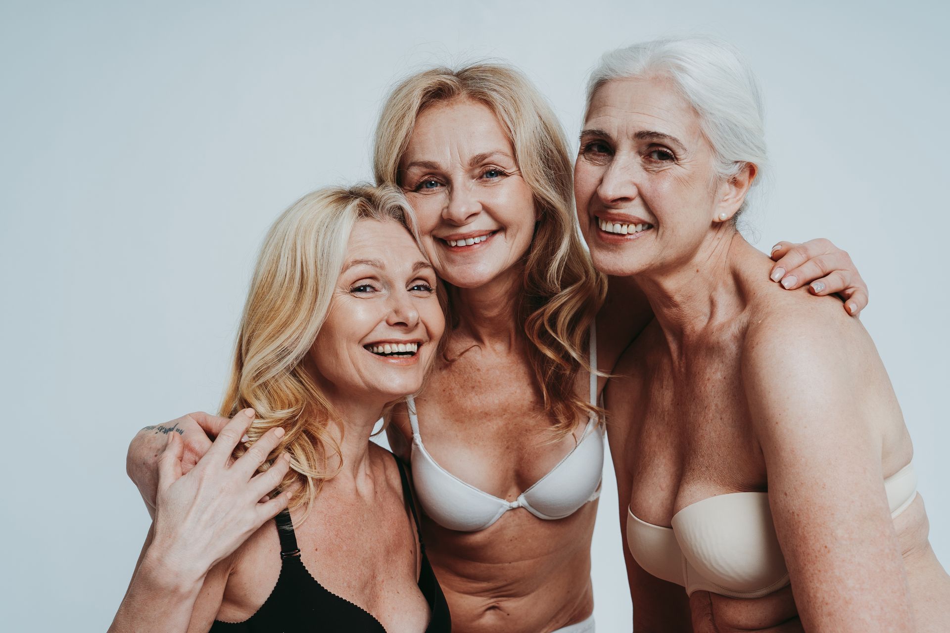 Three smiling women in light-colored lingerie posing closely together against a plain background