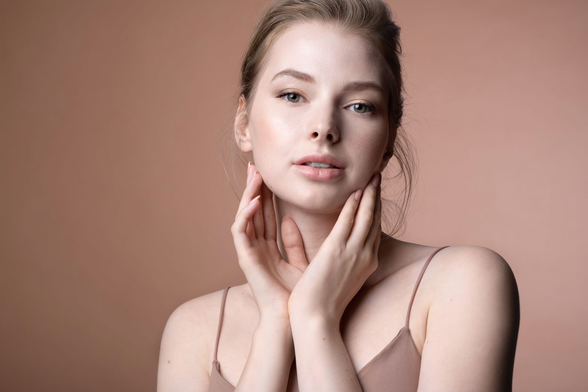 Portrait of a woman in a beige top, resting her hands on her neck against a plain tan background