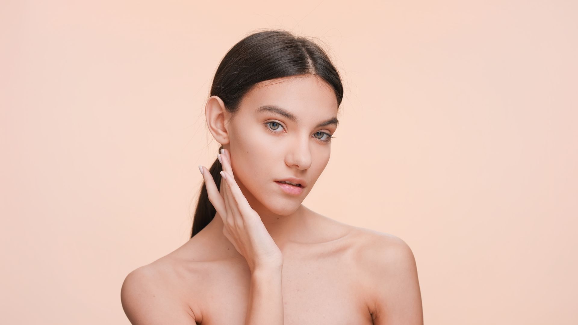 Portrait of a woman against a soft peach background, touching her cheek and looking at the camera.