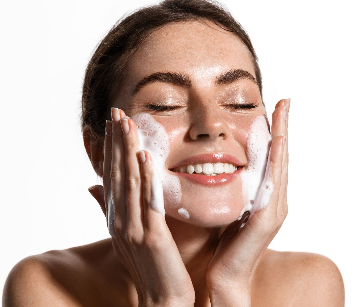 Woman applying facial cleanser to cheeks, smiling with eyes closed against a white background