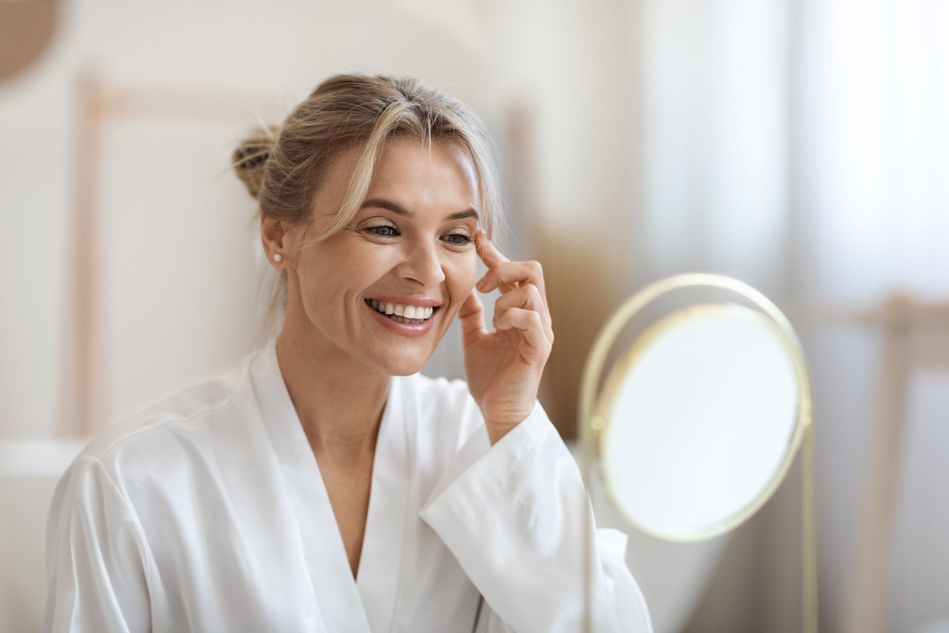 Smiling woman in a white robe applying skincare near a mirror