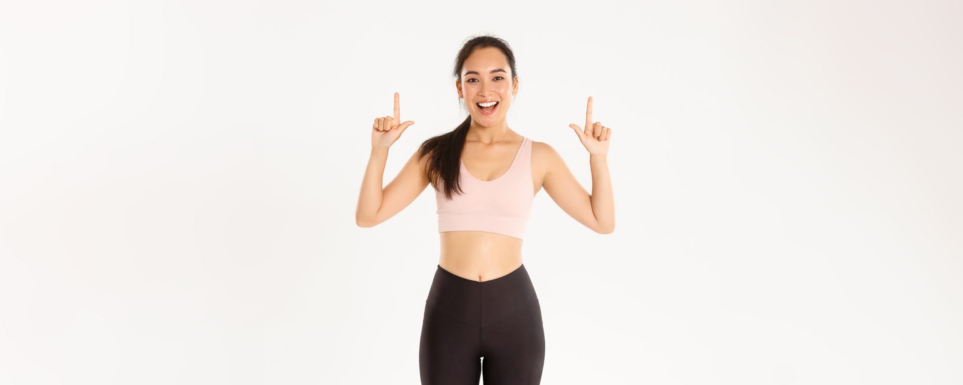 Woman in a beige sports bra and black leggings smiling and pointing upward on a white background