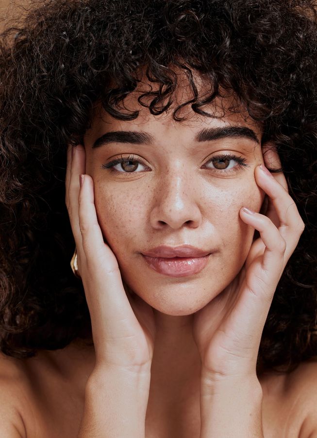Close-up portrait of a person with curly hair, hands on cheeks, looking directly at the camera.