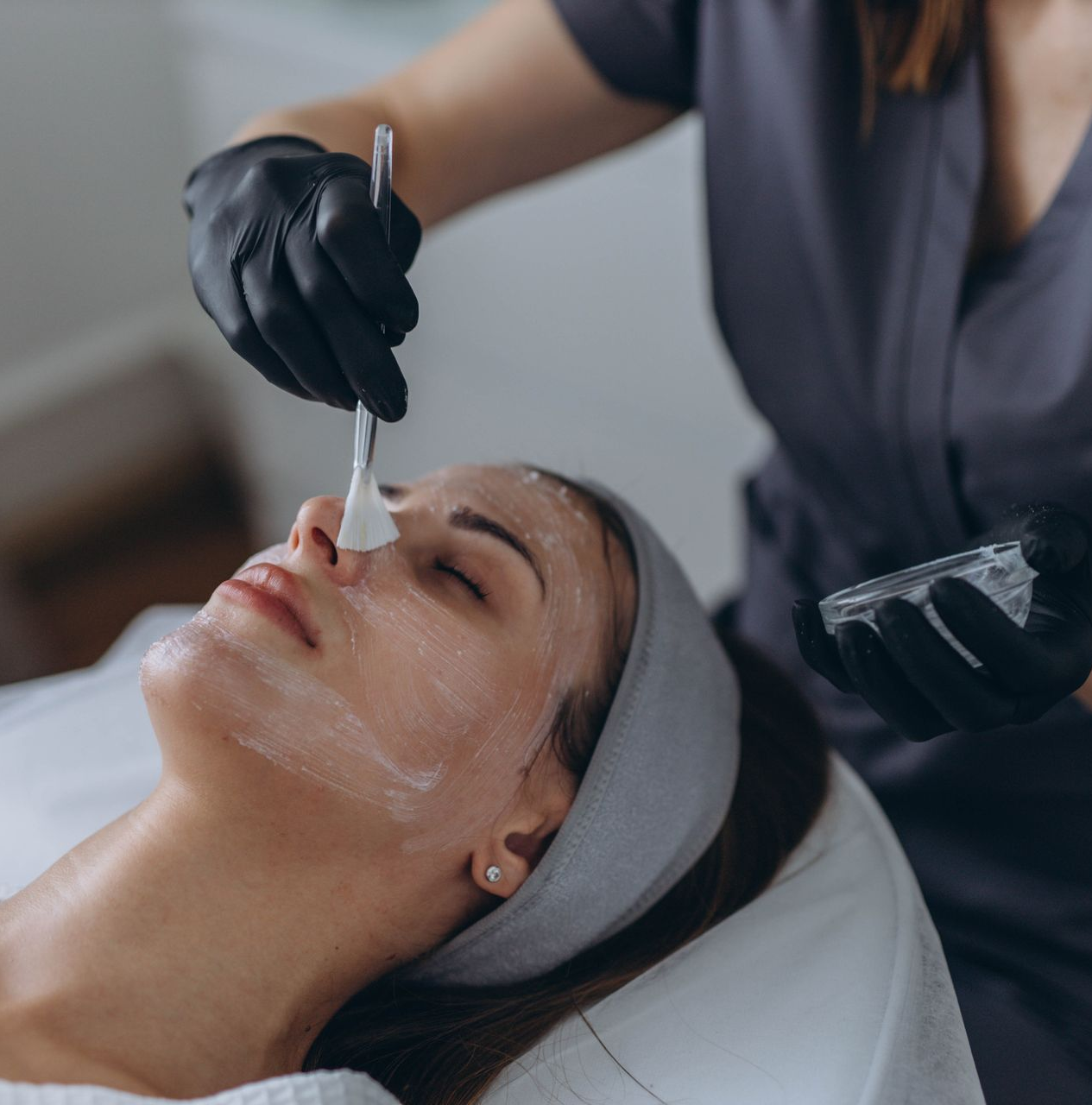 A person receives a facial treatment while a gloved esthetician applies cream with a brush.
