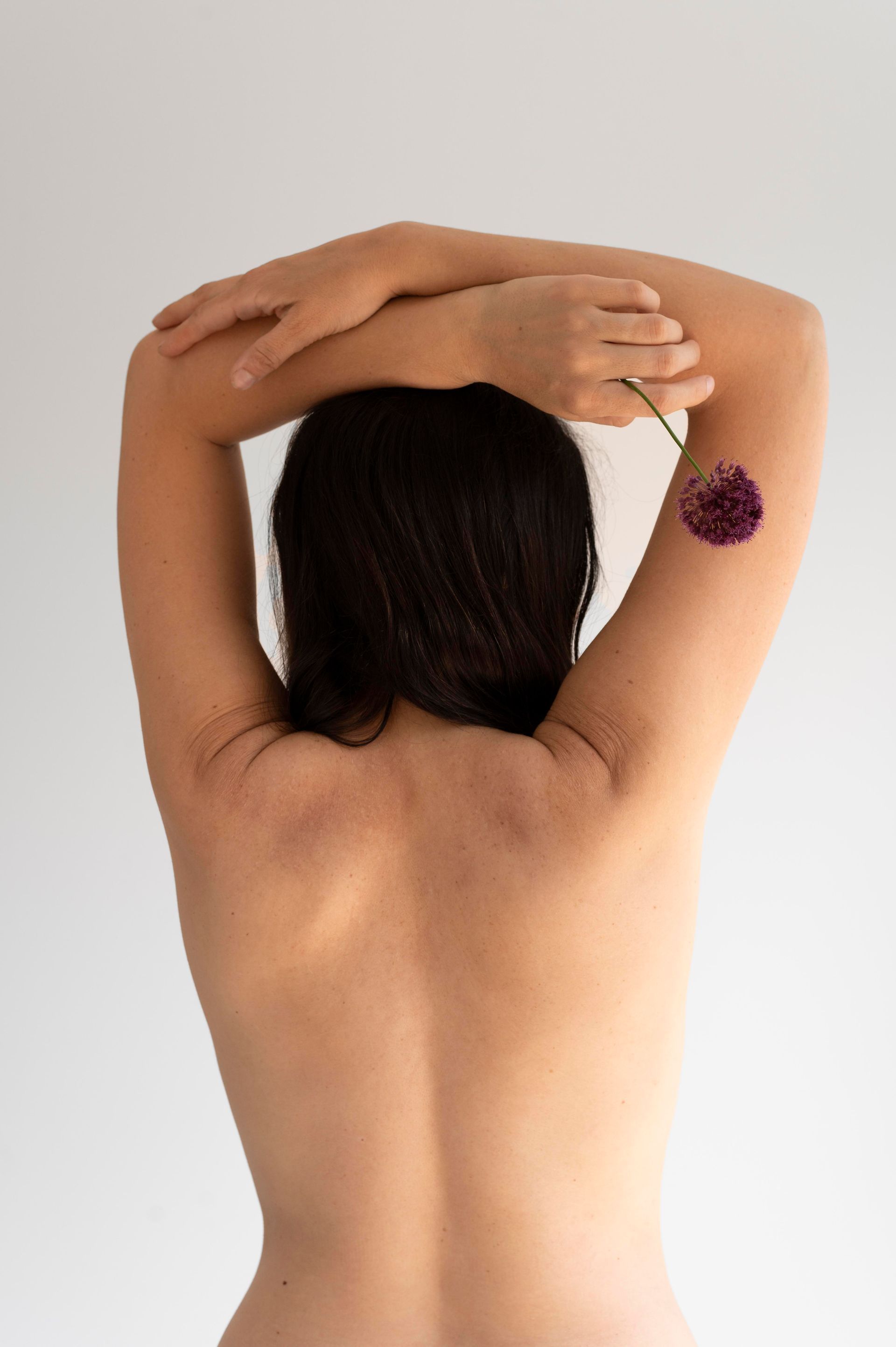 Rear view of a person with arms raised, holding a purple flower against a plain background