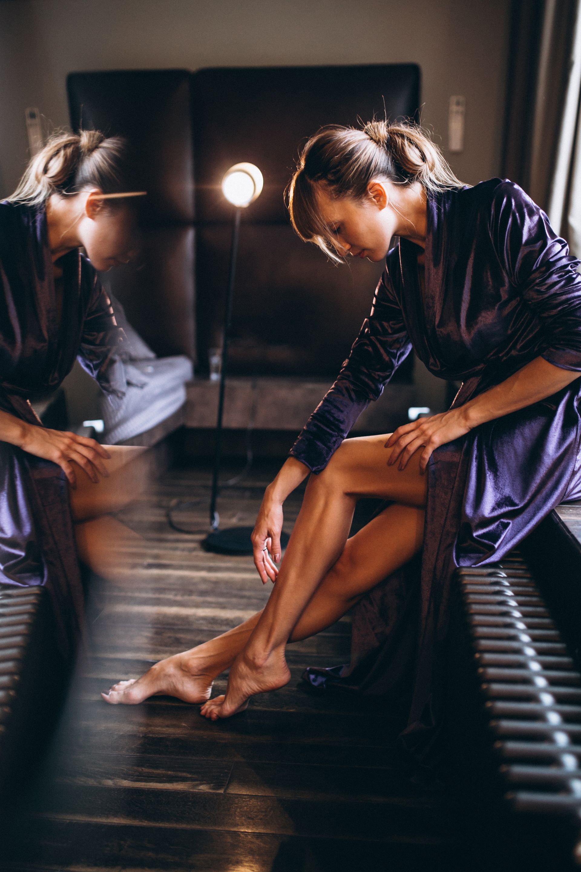 Woman in a satin robe sits on a bench, looking down and adjusting her foot in a dimly lit room.
