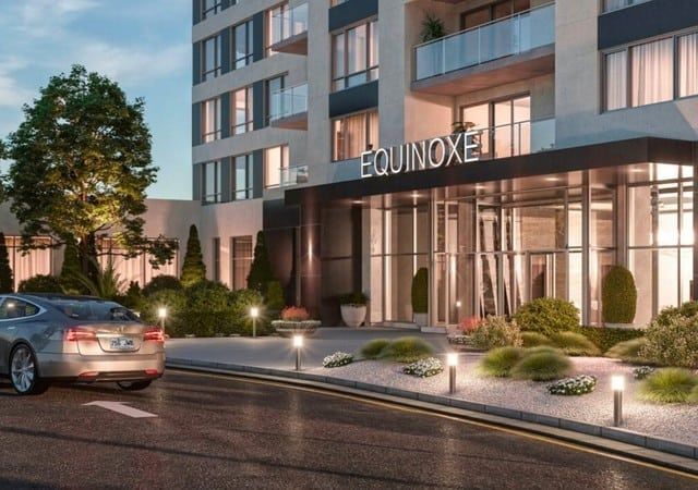 Modern apartment building entrance at dusk with lit glass lobby, parked car, and “EQUINOXE” sign
