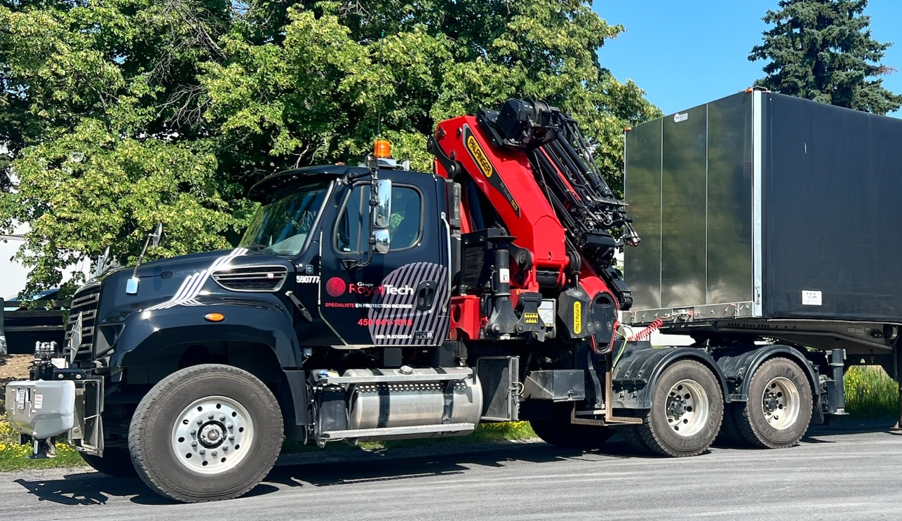 Un semi-remorque noir avec une grue rouge transporte un grand conteneur sombre sur une route sous un ciel bleu.
