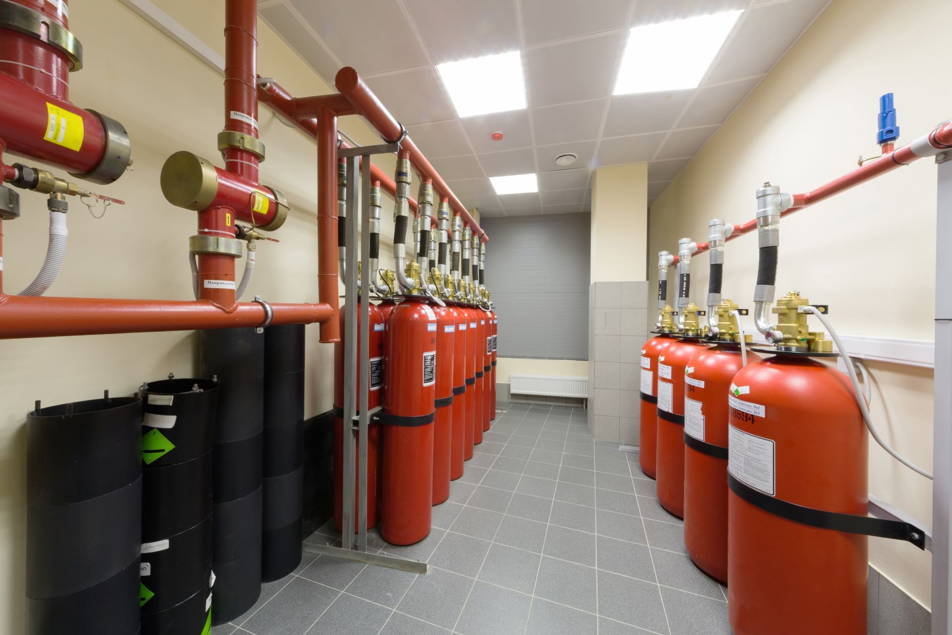 Local technique d'extinction d'incendie avec bouteilles rouges, réservoirs noirs et canalisations aériennes dans un couloir carrelé