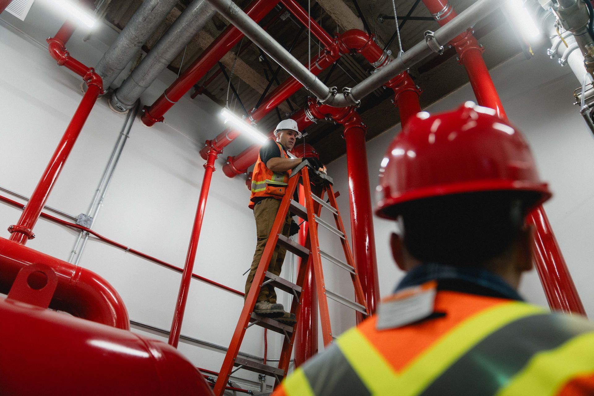 Des ouvriers portant des casques de chantier inspectent des tuyauteries industrielles rouges dans un local technique, l'un d'eux se trouvant sur une échelle.