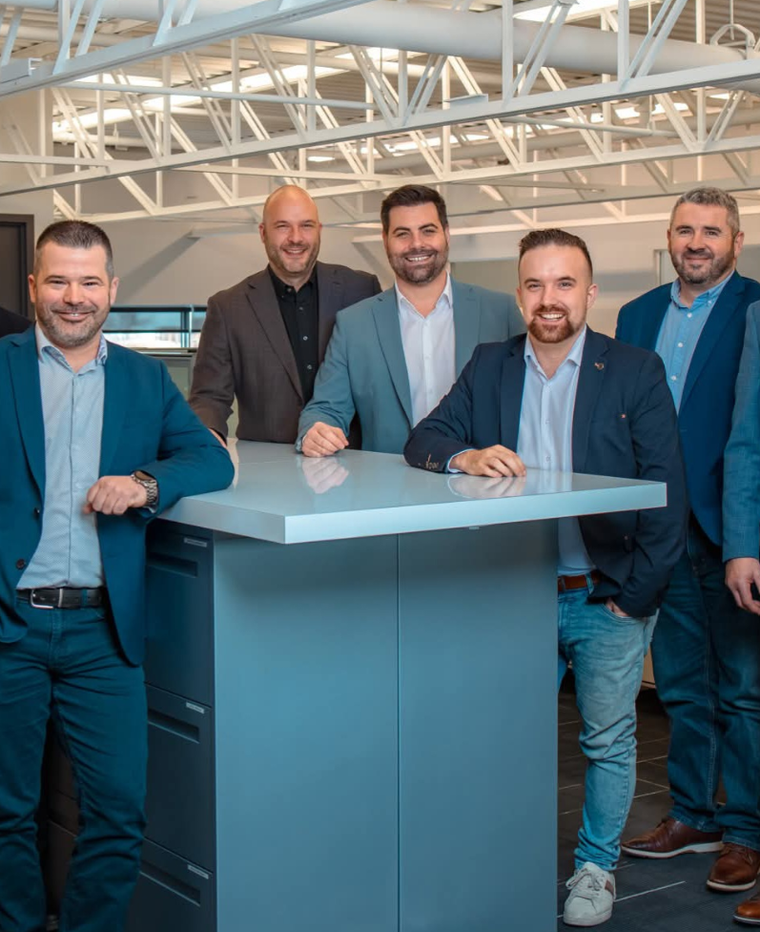 Cinq hommes en costume bleu se tiennent debout autour d'une table haute turquoise dans un bureau lumineux.