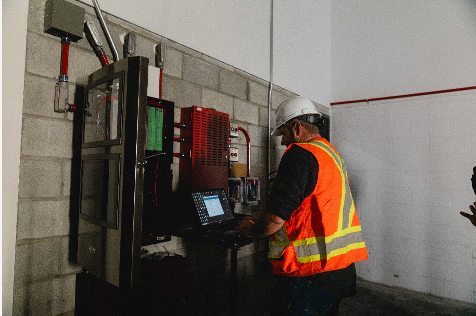 Ouvrier portant un gilet de sécurité orange et un casque de chantier manipulant un panneau de commande dans une salle industrielle