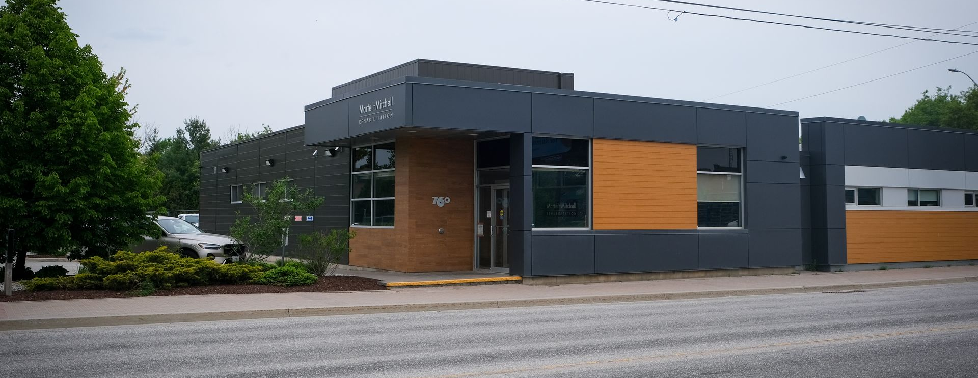 A modern, flat-roofed commercial building with grey panels and orange accents, set near a road with trees nearby.