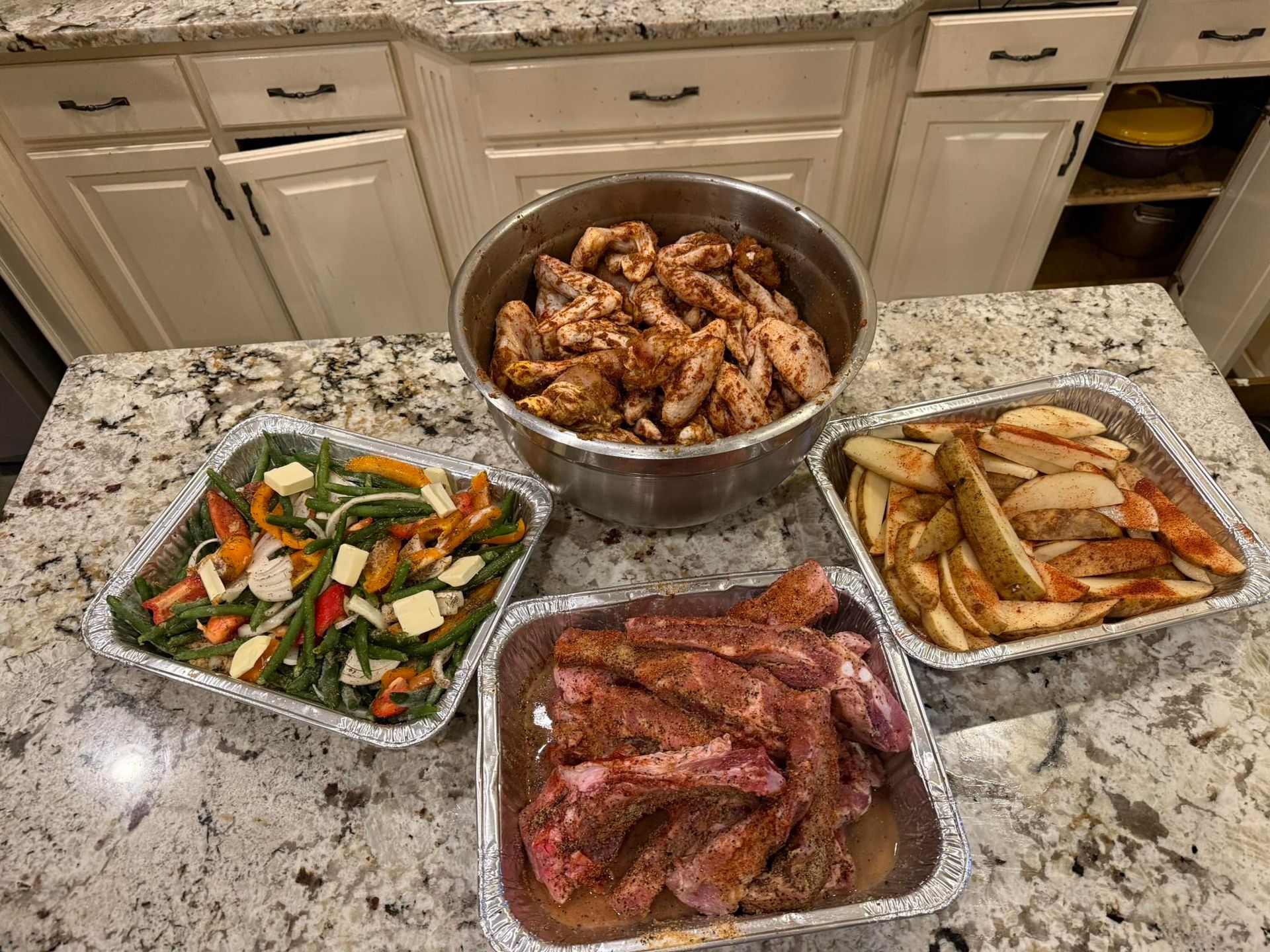 Chicken wings, ribs, potato wedges, and vegetables in foil pans on a countertop.