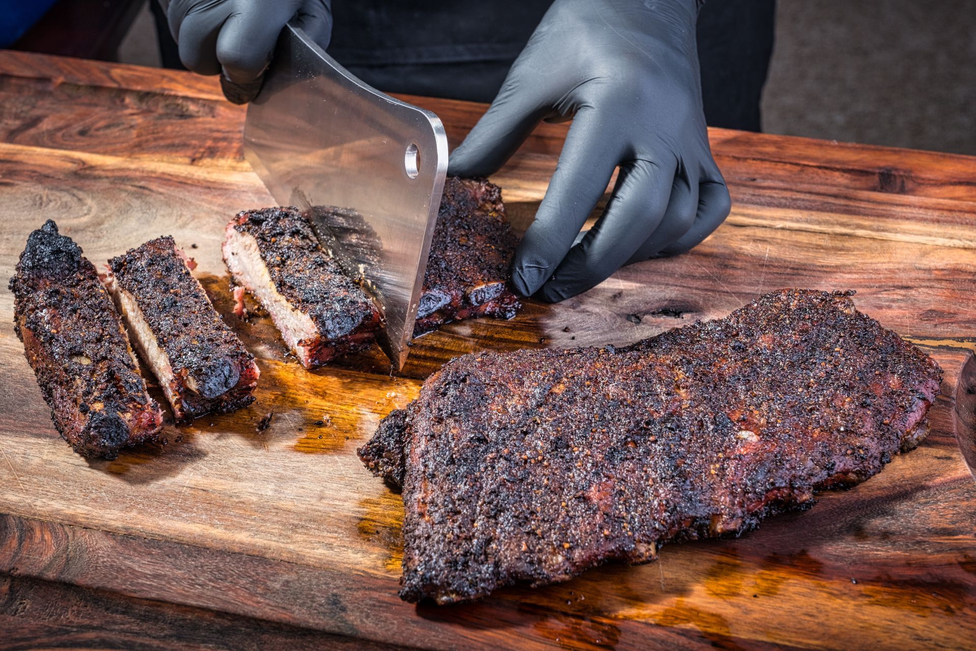 Person in black gloves slicing smoked ribs on a wooden cutting board with a cleaver. Person in black gloves slicing smoked ribs on a wooden cutting board with a cleaver.