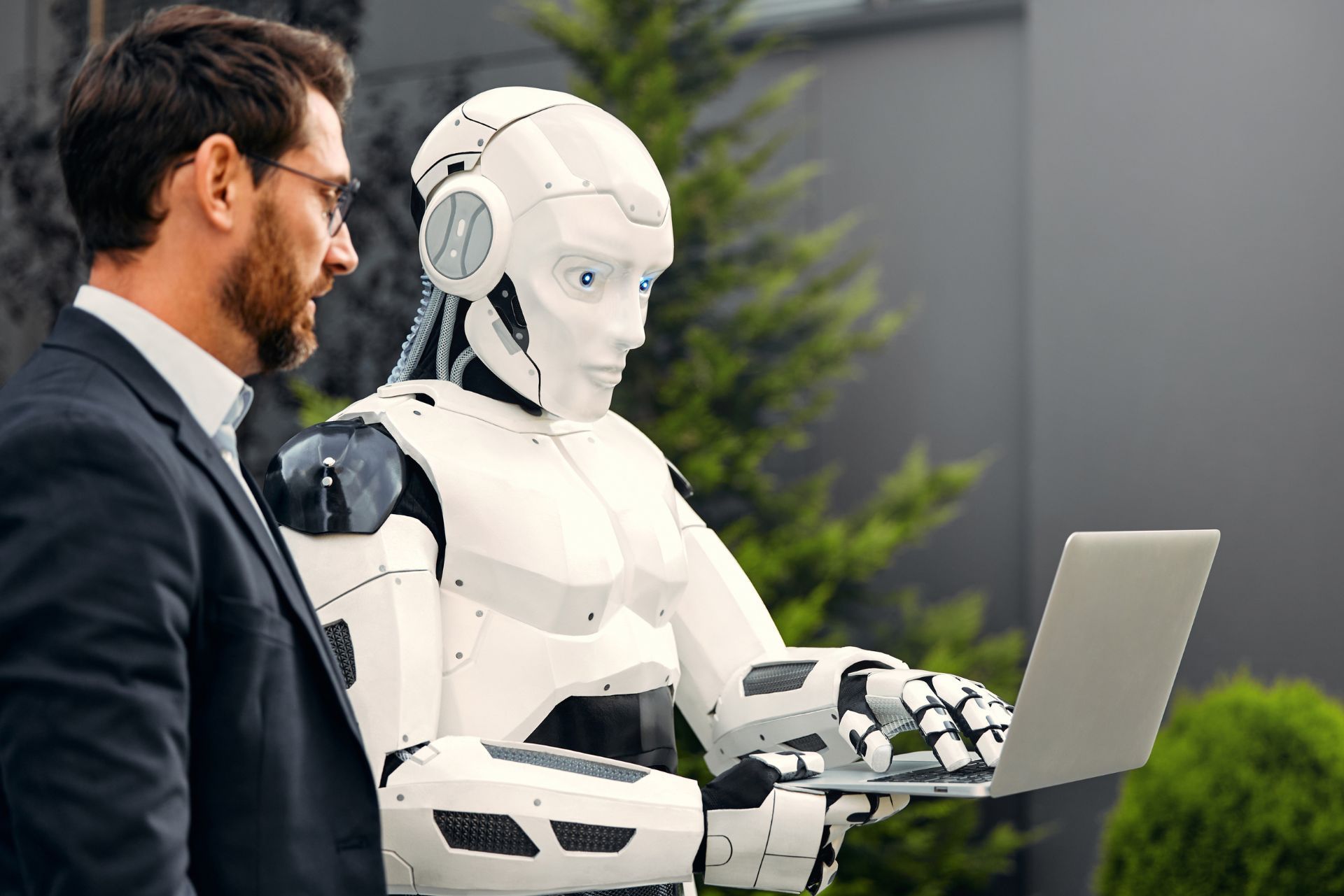 A human in a suit stands next to a humanoid robot as it types on a laptop outdoors.