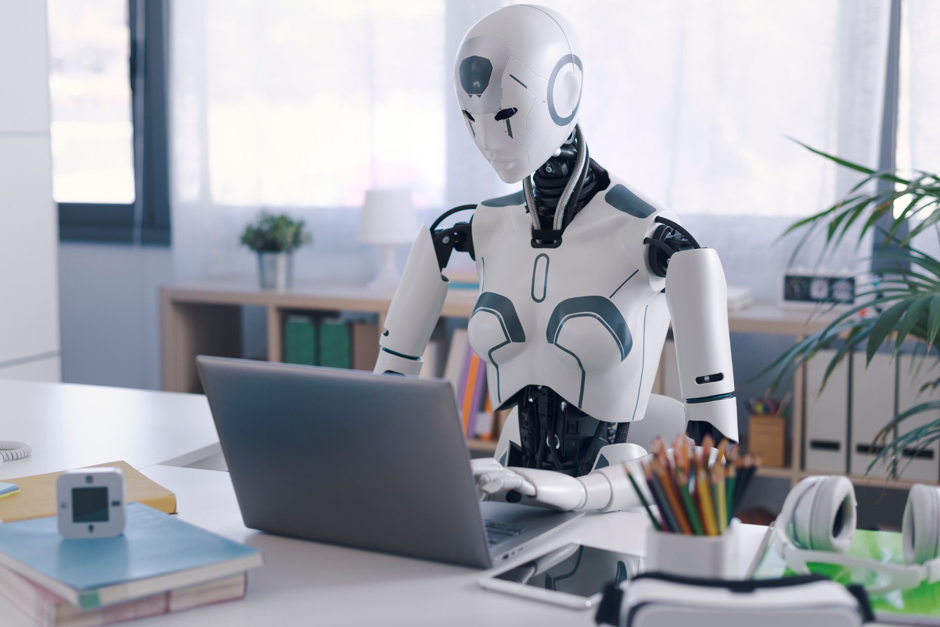 A white humanoid robot typing on a laptop at a desk in a bright office.