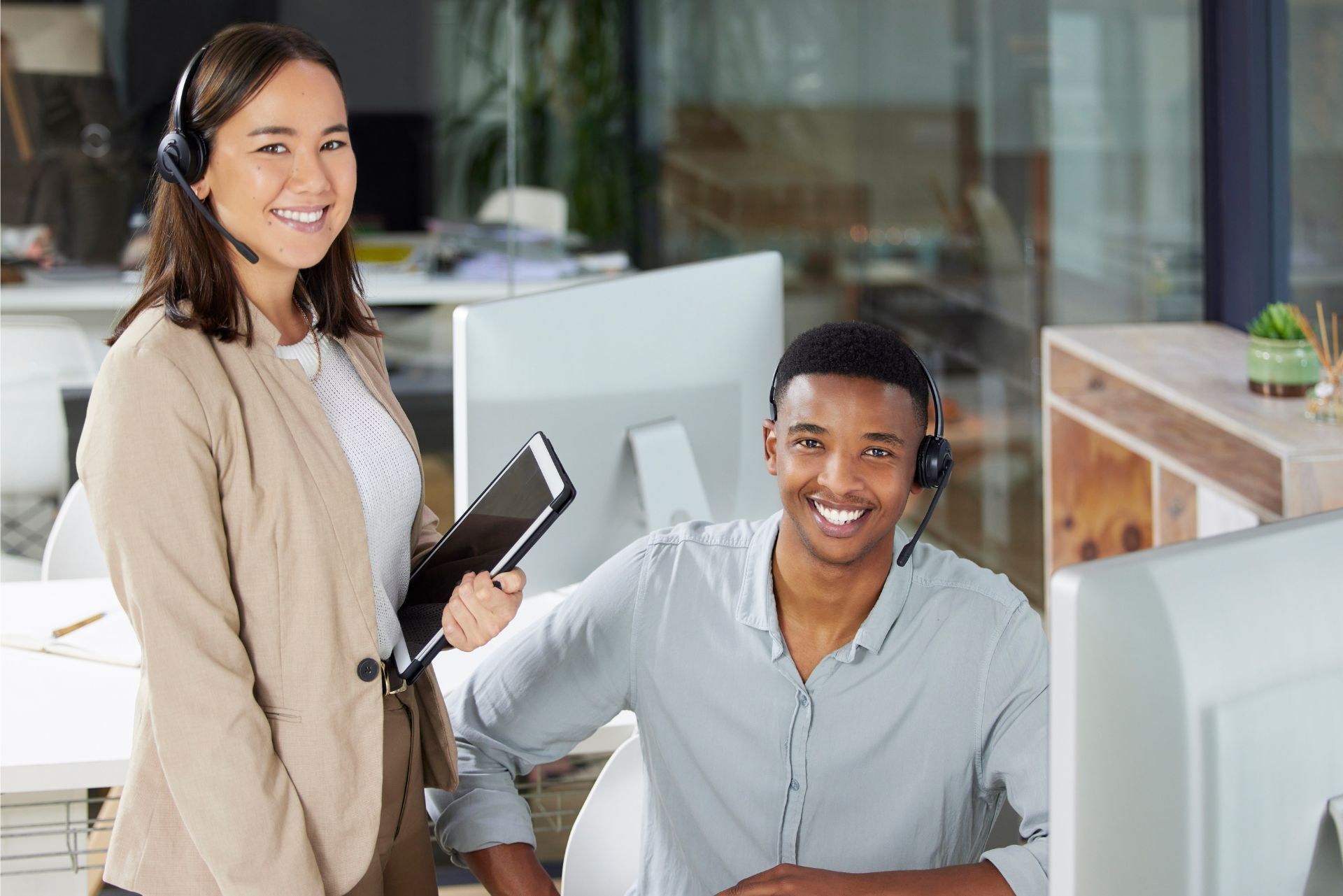 Two coworkers smiling in a bright office, one standing with a tablet beside a seated colleague at a desk.