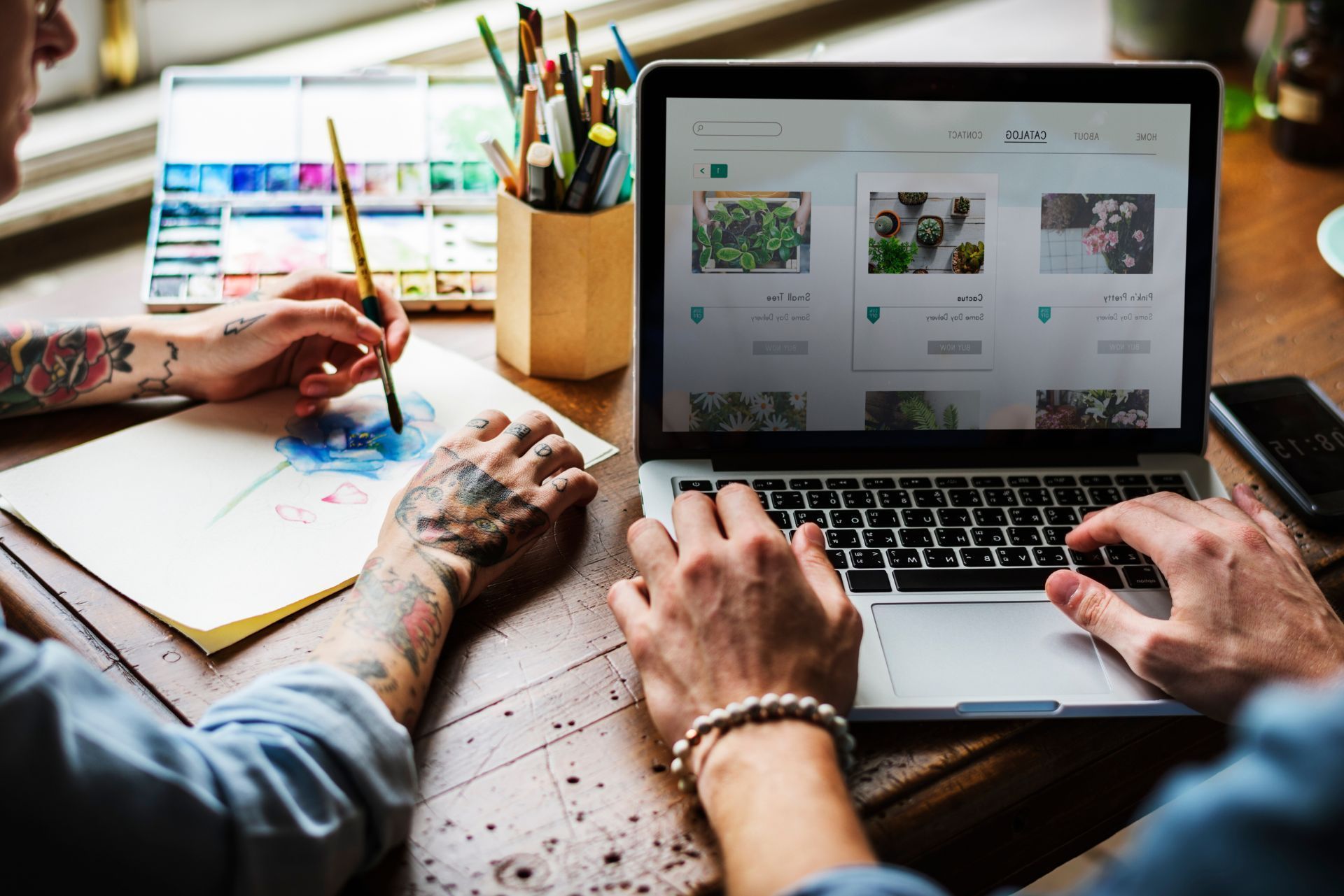 Two people working at a desk, one writing on paper and one using a laptop with design thumbnails onscreen