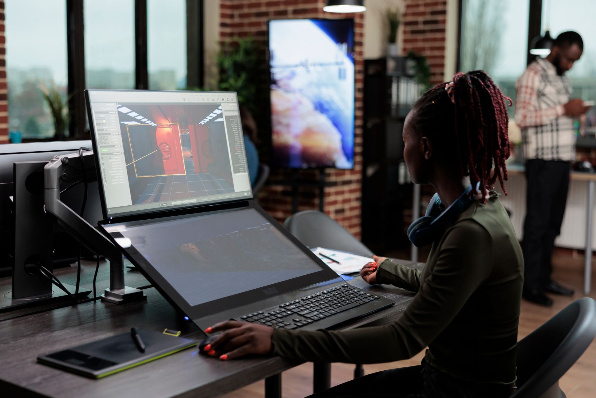 Person using a laptop and monitor at a desk in a dimly lit office, with a city view and another person in the background