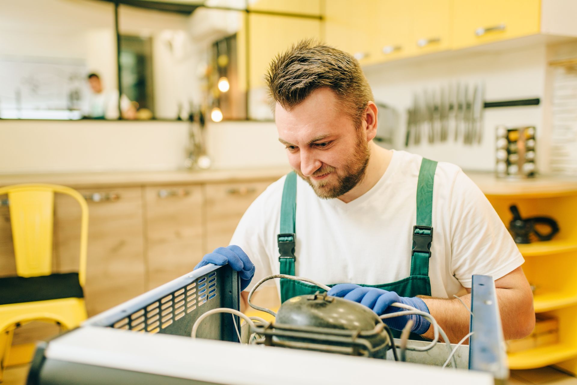 Man in green overalls repairing a dishwasher in a bright kitchen repair shop