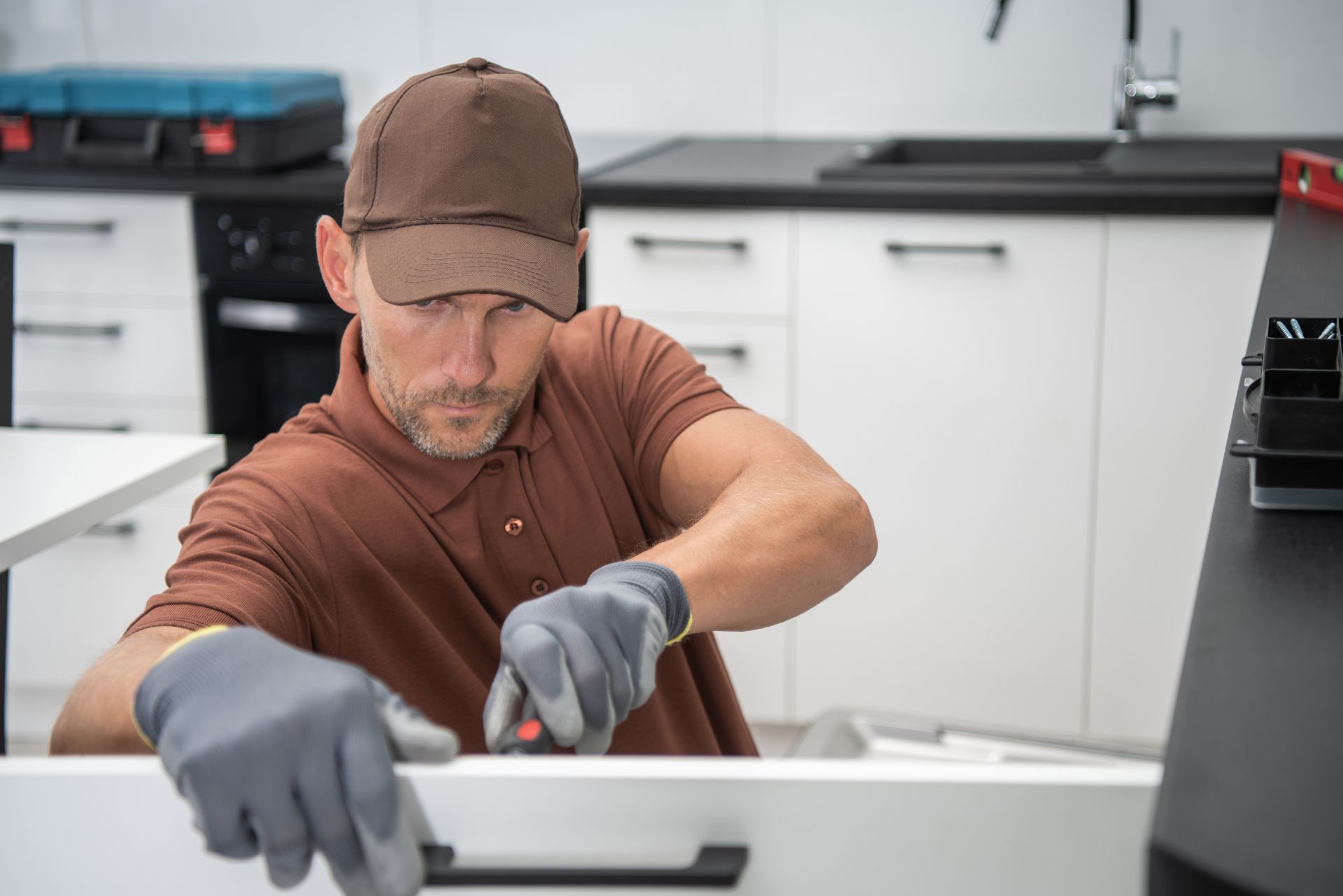 Worker installing a drawer in a modern kitchen, wearing gloves and a cap.