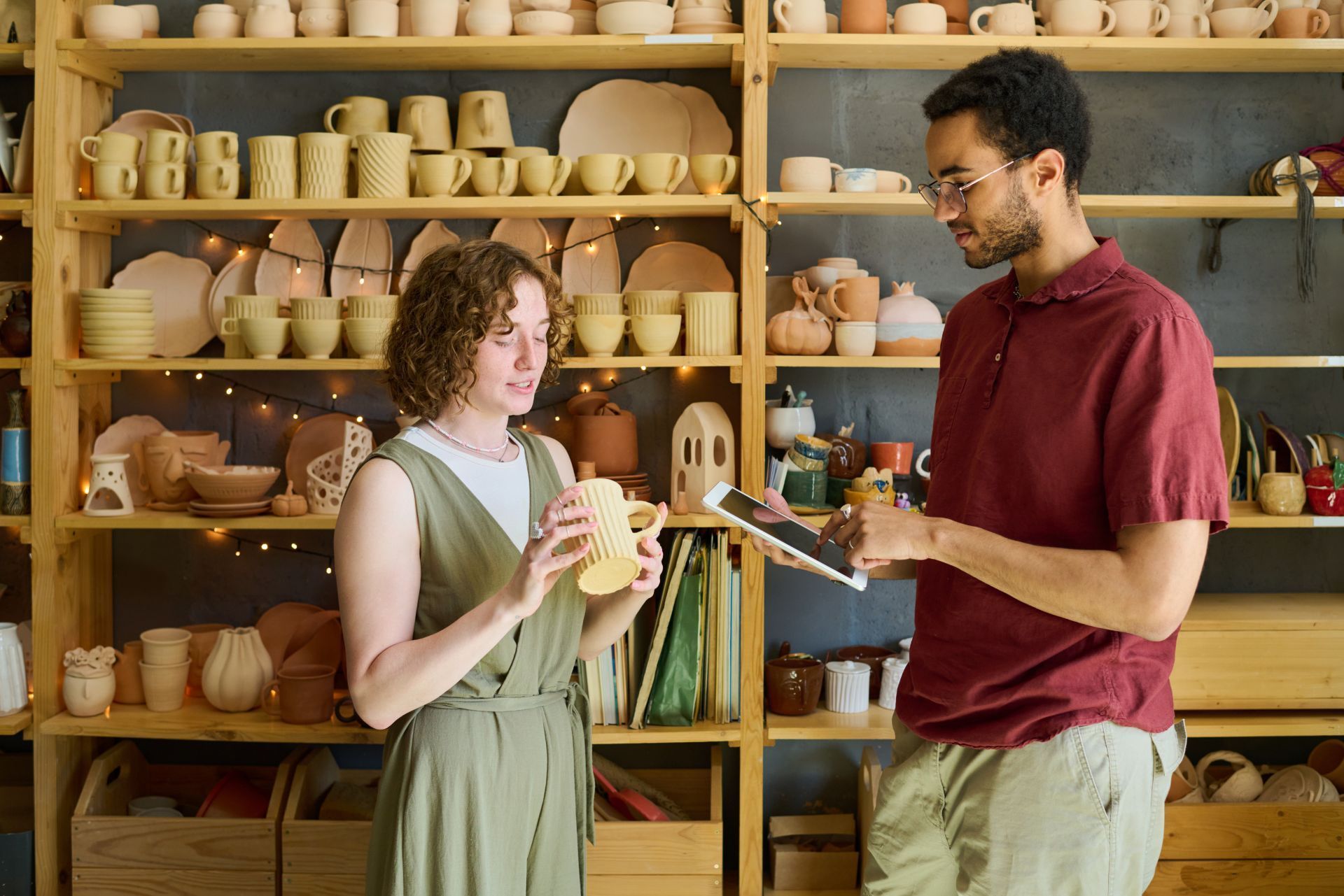 Two people discussing a pottery piece in a ceramics studio lined with shelves of handmade mugs and bowls