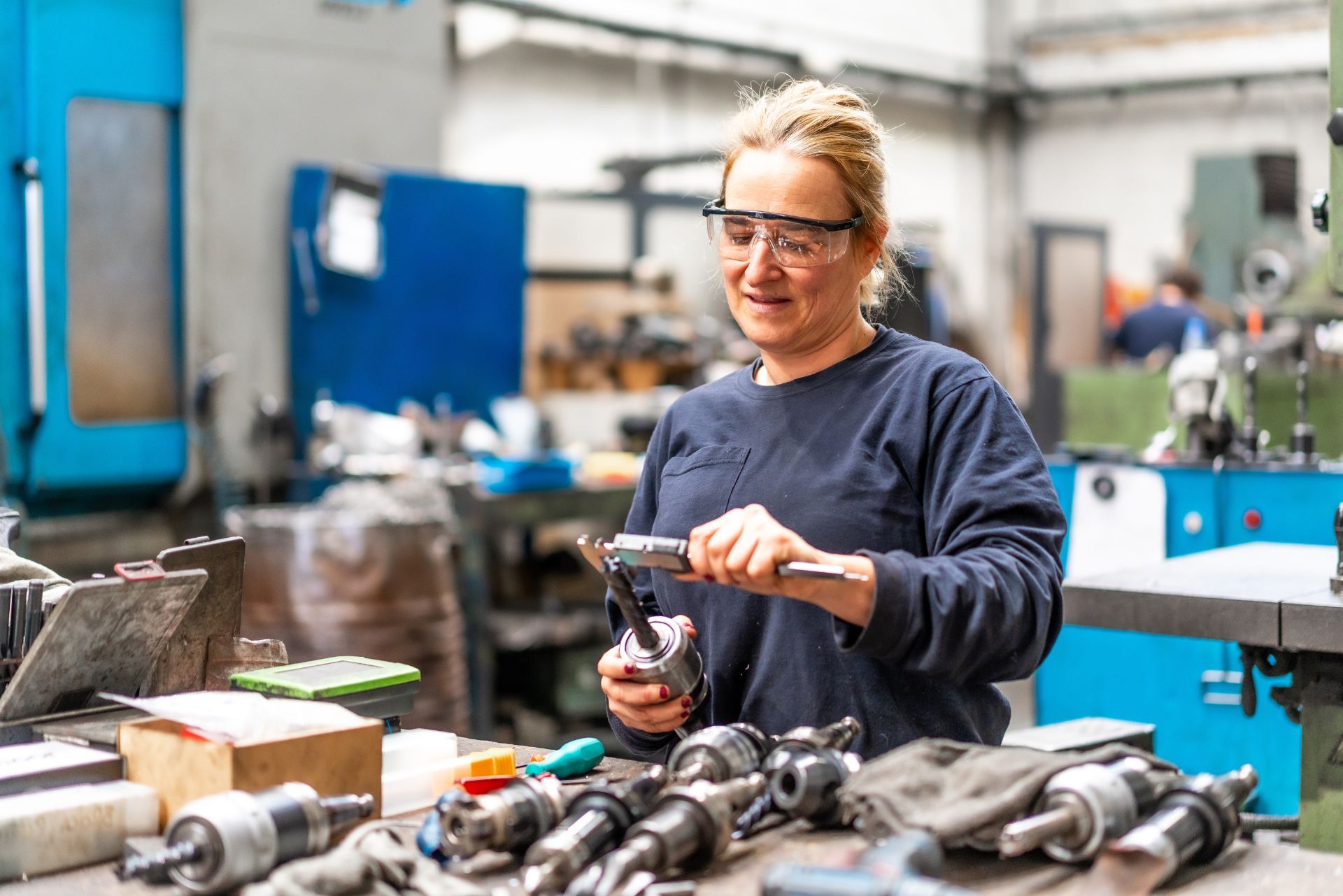 Worker in safety glasses using a hand tool at a metal workbench in a machine shop.