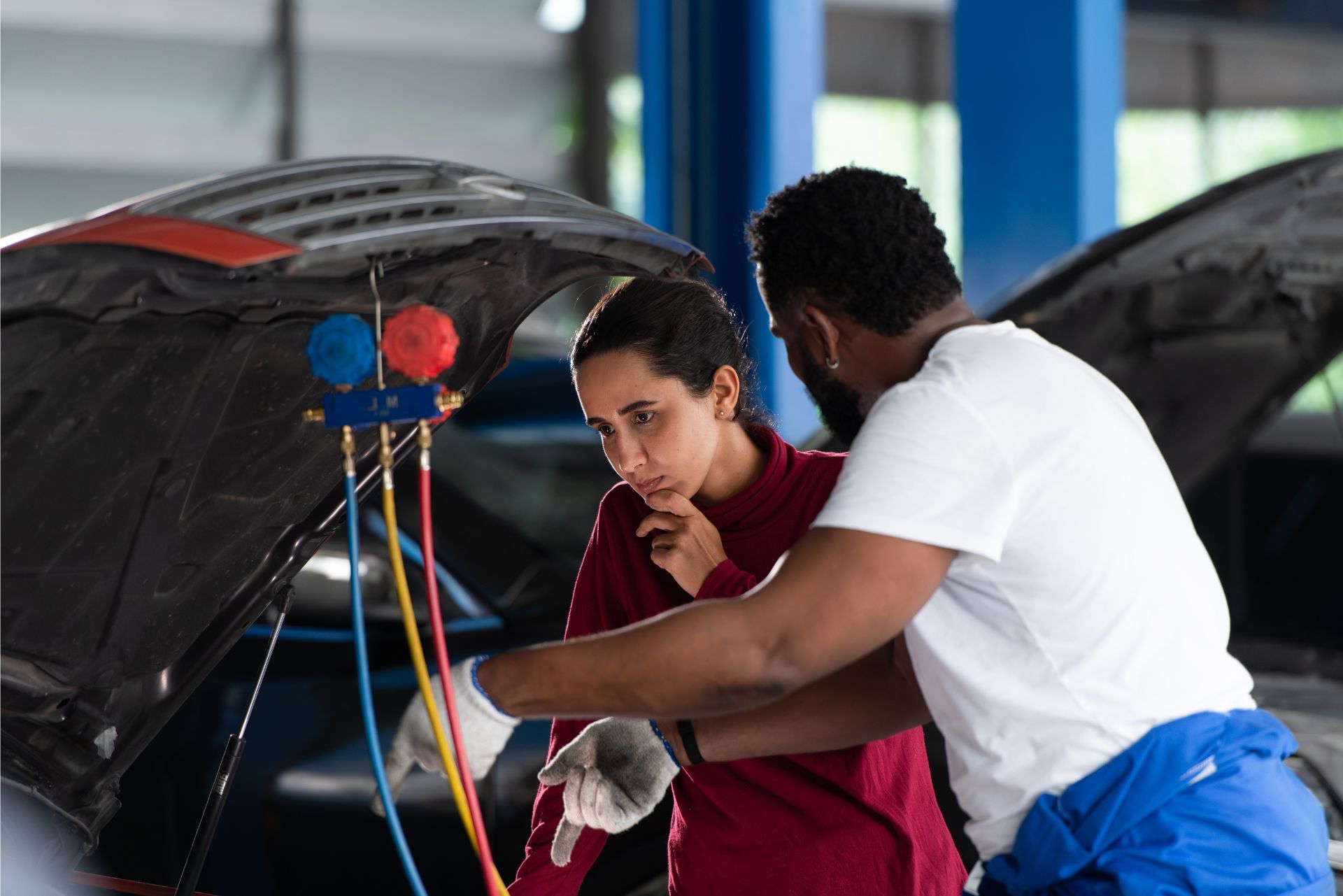 Two people inspect a car engine with the hood open, one using tools in a garage.