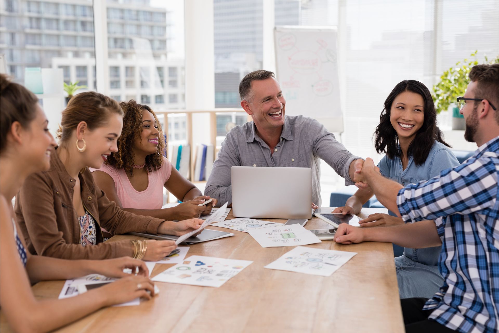 A diverse group of colleagues smiles while sitting around a wooden table in a bright office, with two people shaking hands.