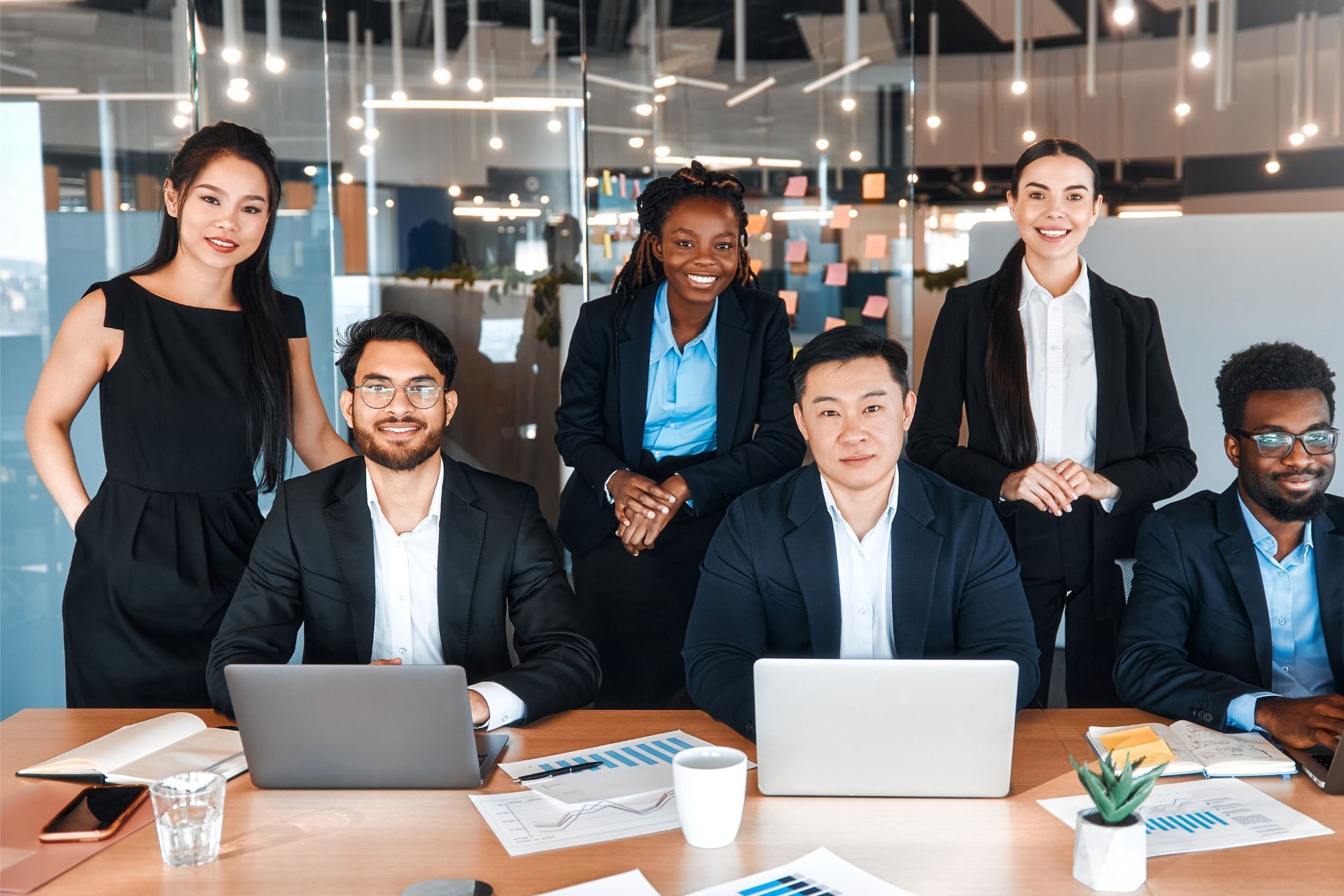 A diverse group of six professionals in business attire smile in a bright, modern office with laptops on a wooden table.