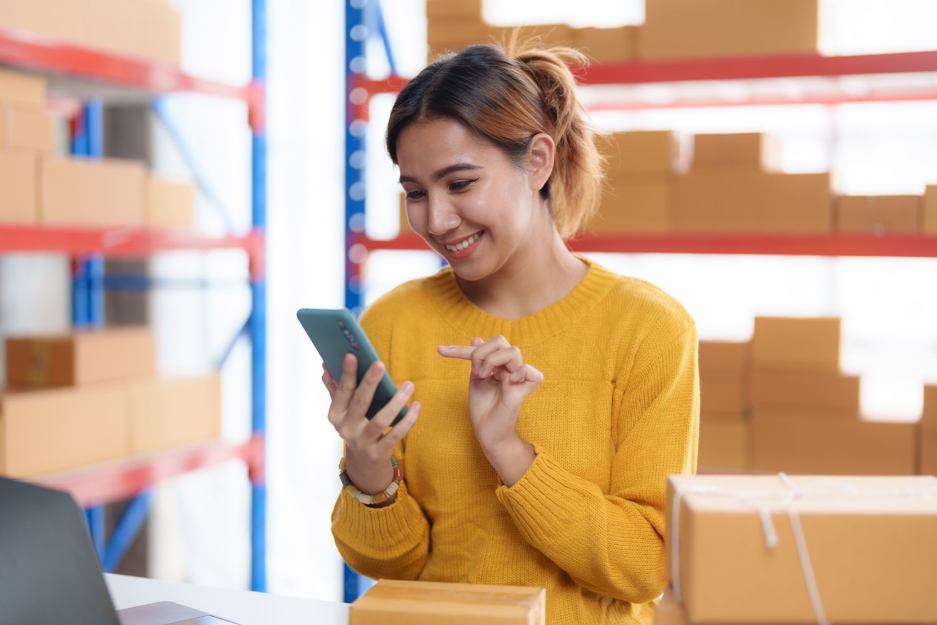 Woman in a yellow sweater smiling at her phone in a warehouse with stacked boxes