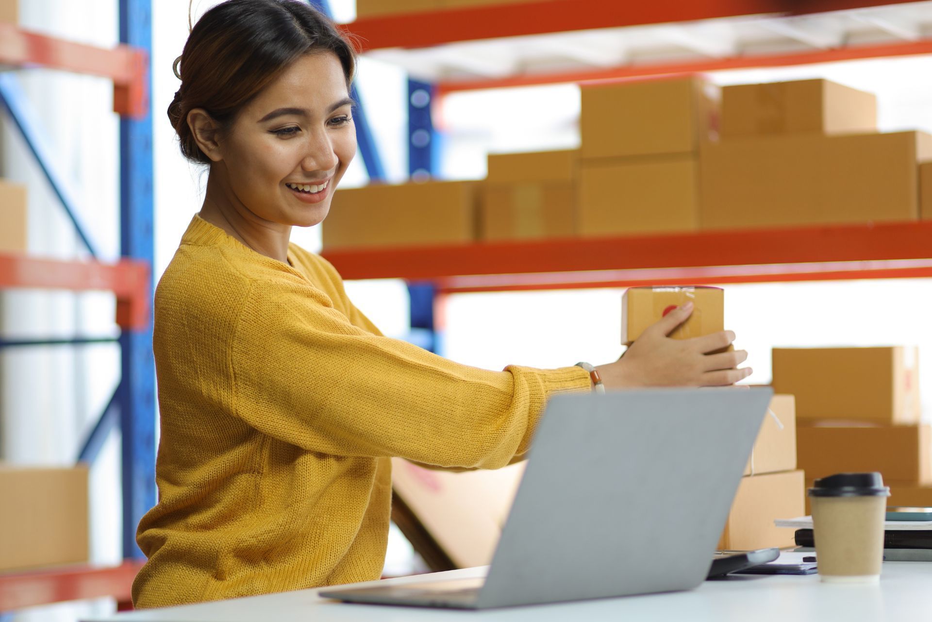 Person packing a small box at a desk with a laptop in a warehouse setting