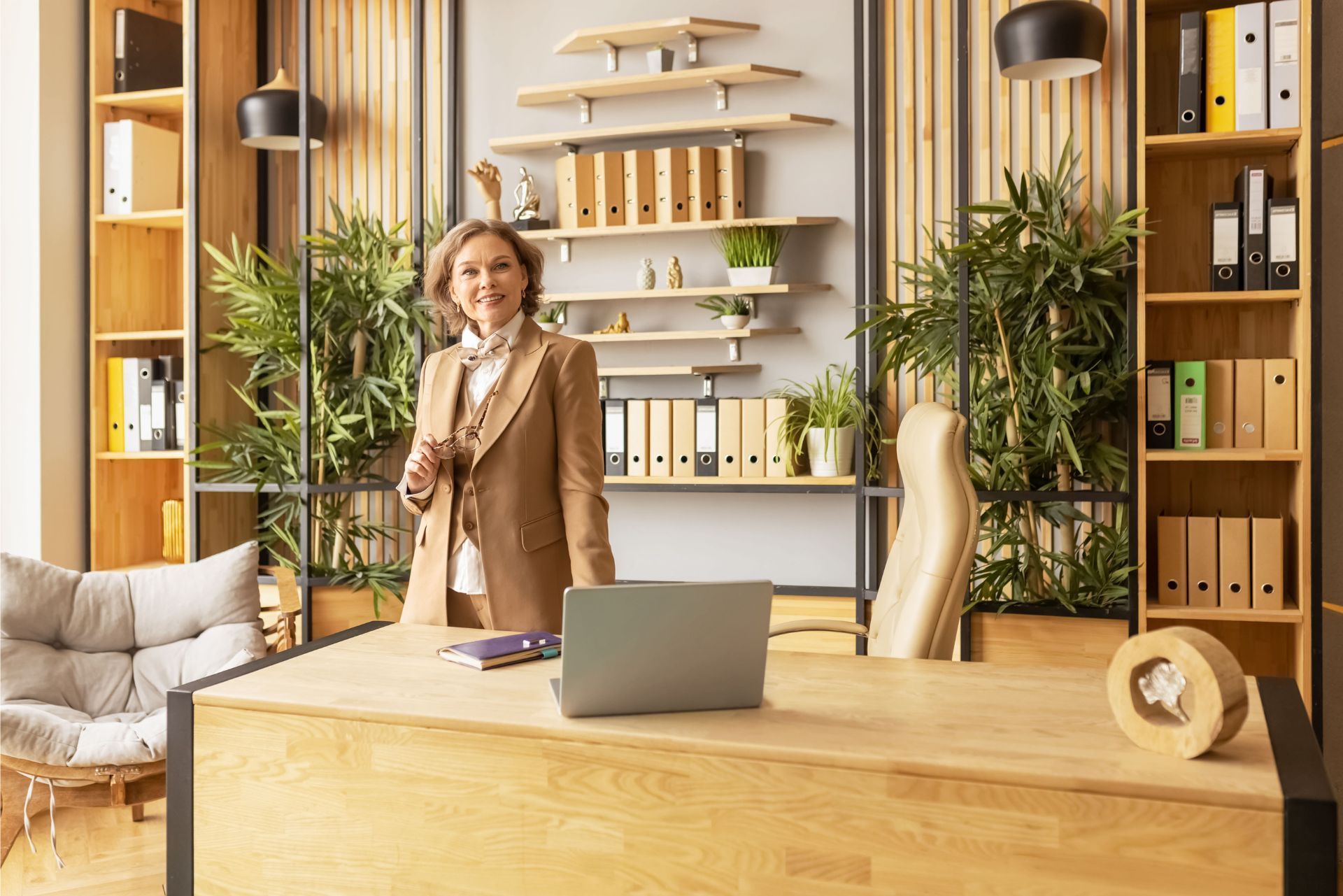 Woman in a bright office behind a wooden desk, with a laptop, shelves of files, and potted plants.