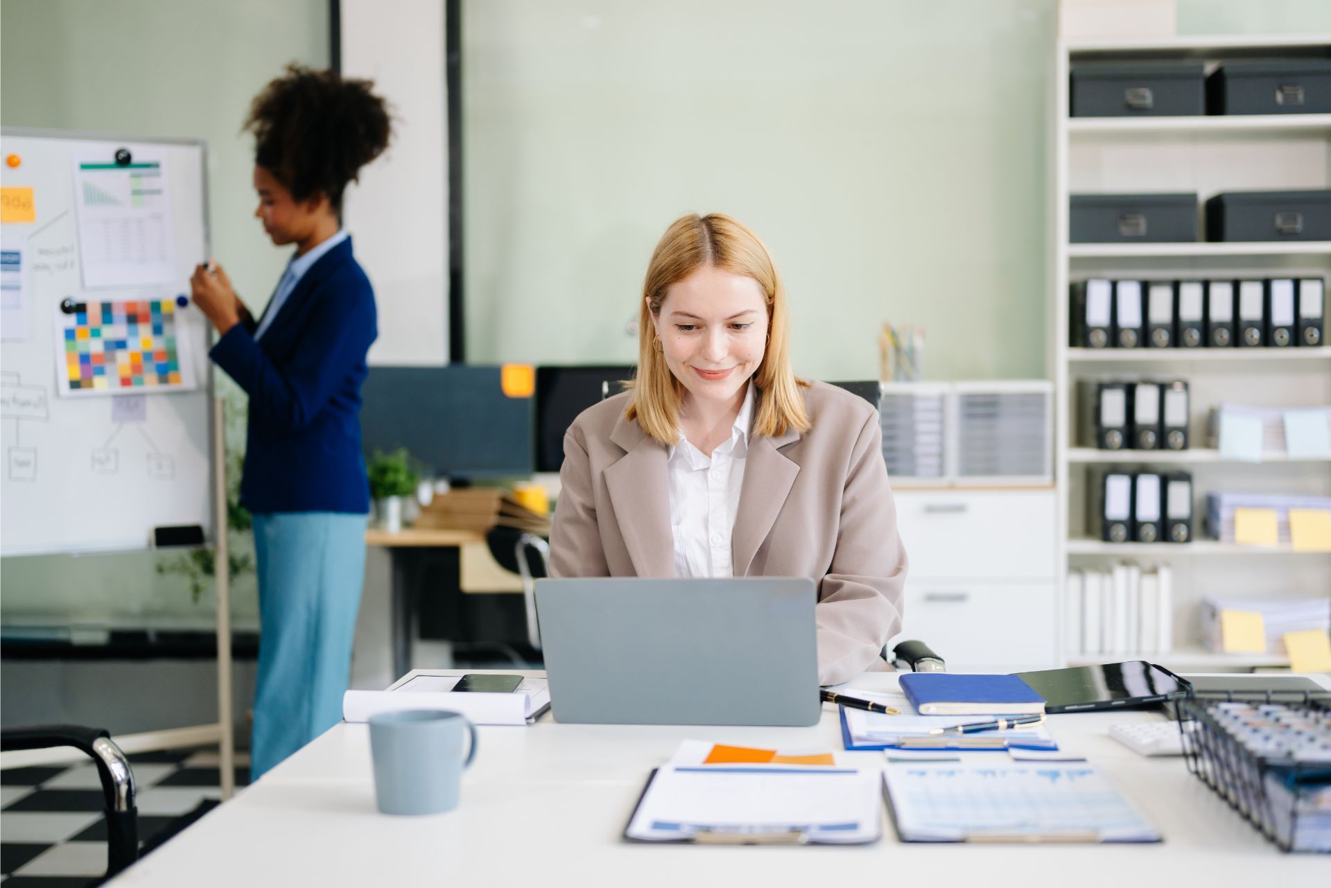 Woman working on a laptop at a desk in a bright office while a colleague presents at a whiteboard.