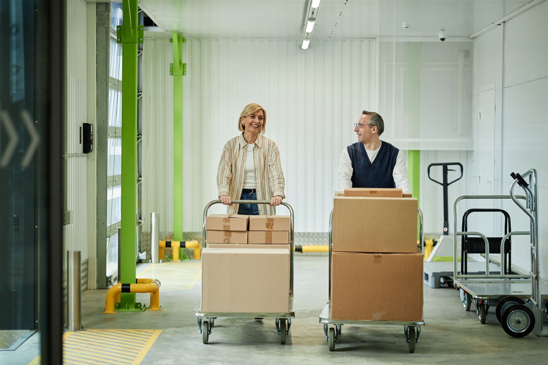 Two workers push loaded carts through a bright warehouse aisle