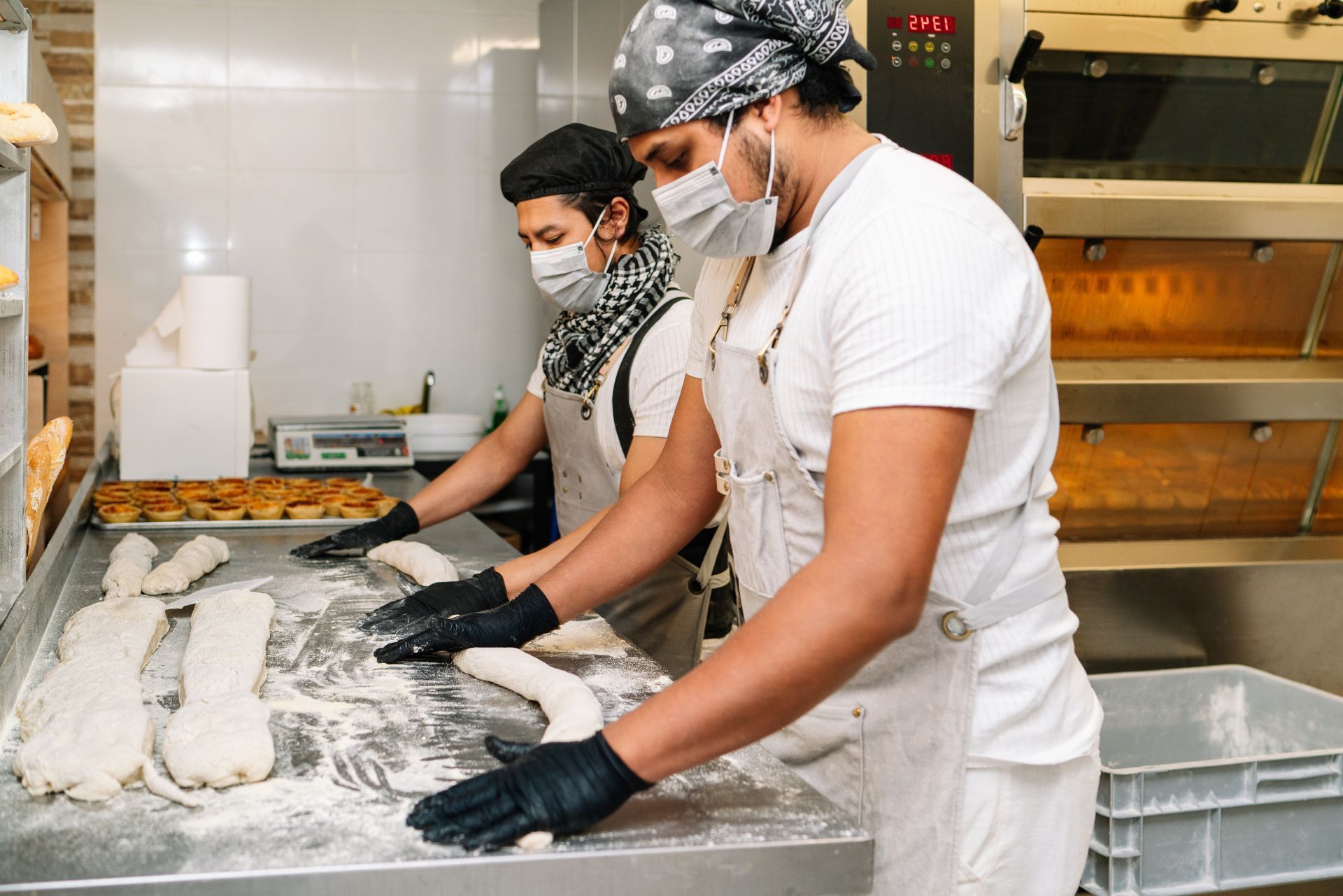 Two bakers in masks, aprons, and gloves knead dough on a flour-dusted stainless steel table in a professional kitchen.