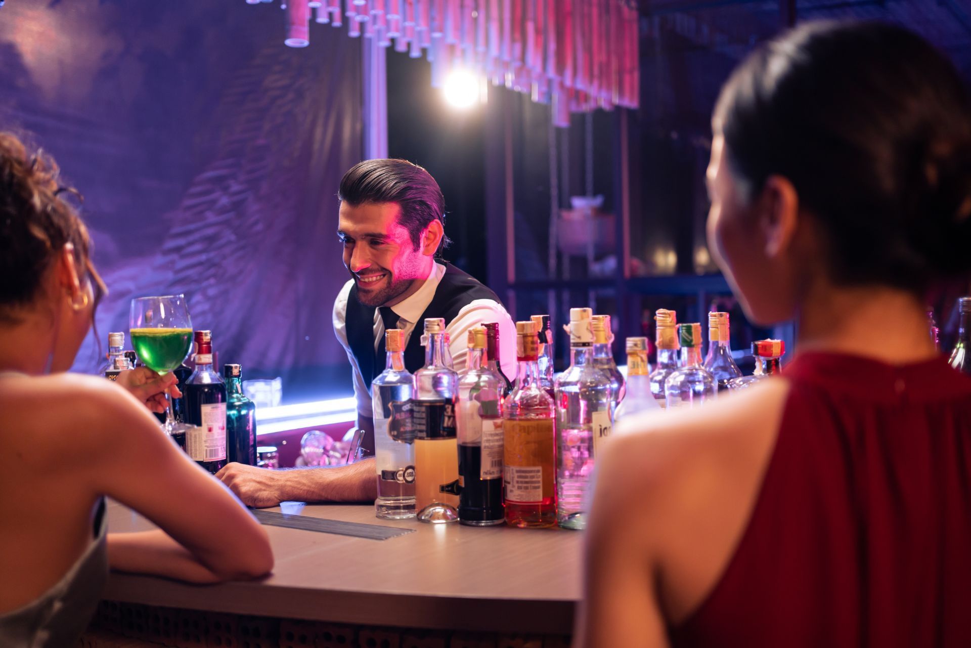 People socializing at a bar, with a smiling host behind a table of drinks and bottles.