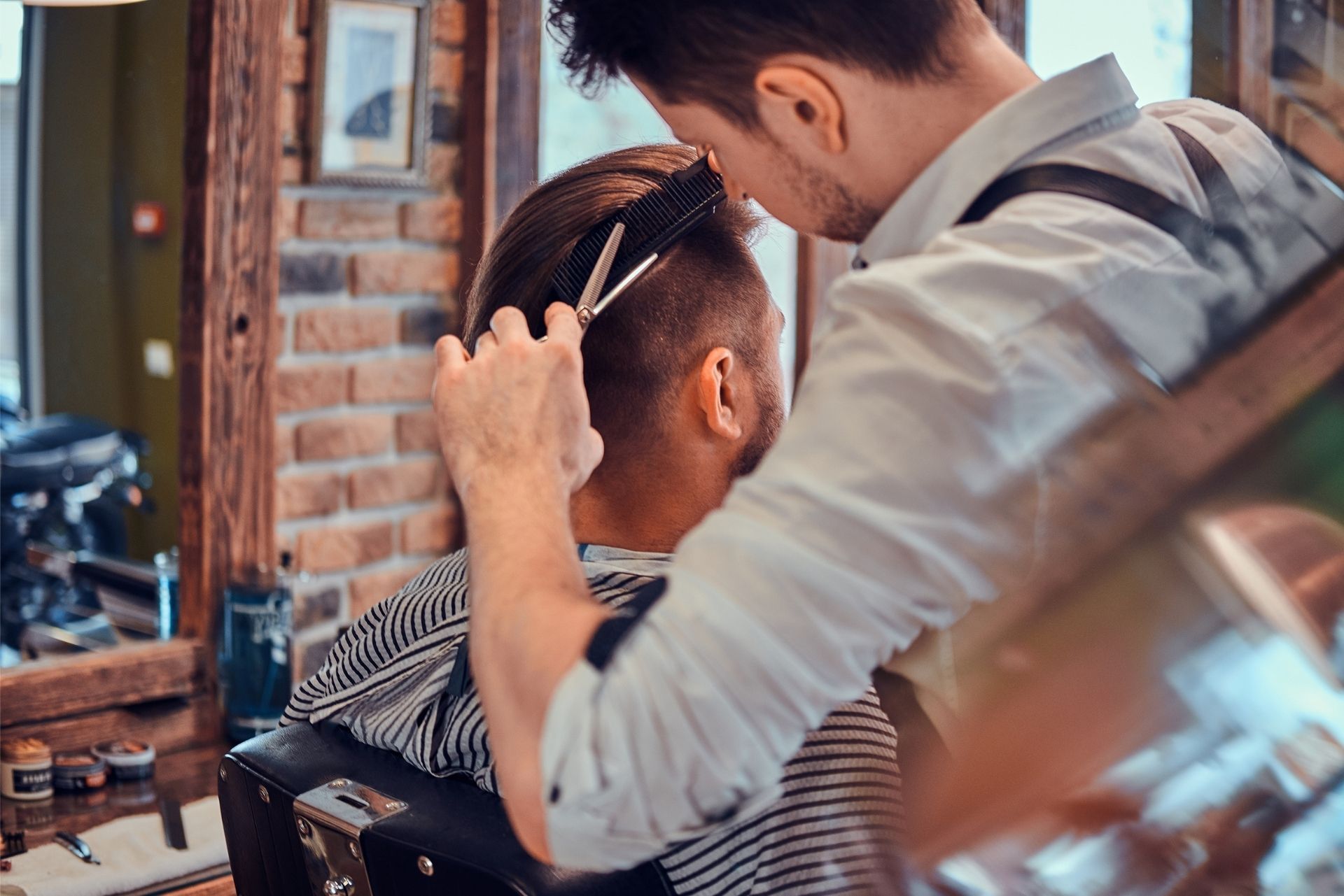 A barber in a light shirt uses scissors and a comb to trim a client's hair in a salon with a brick wall.