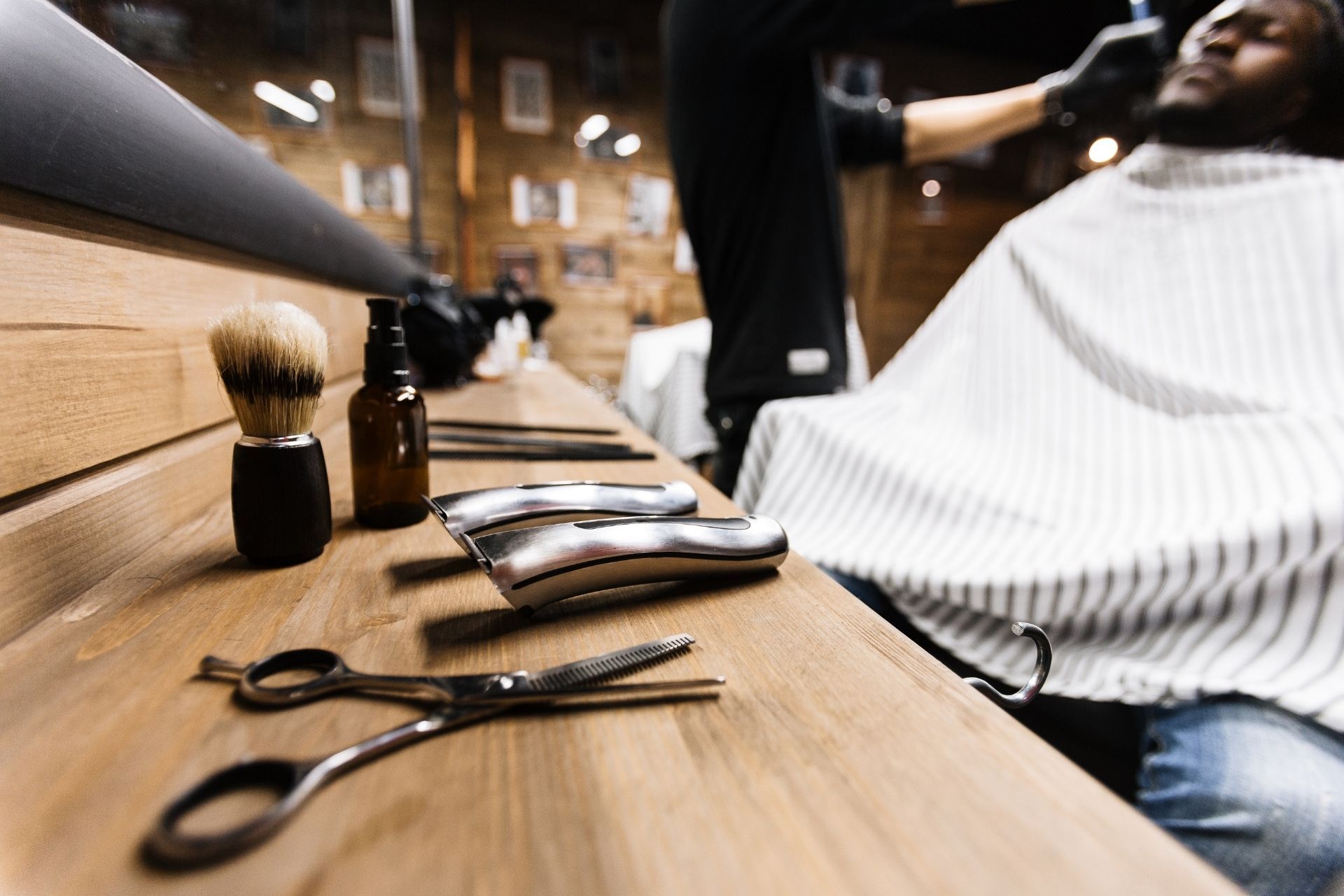 Barber tools, including scissors, clippers, and a brush, rest on a wooden counter as a barber works in a shop.