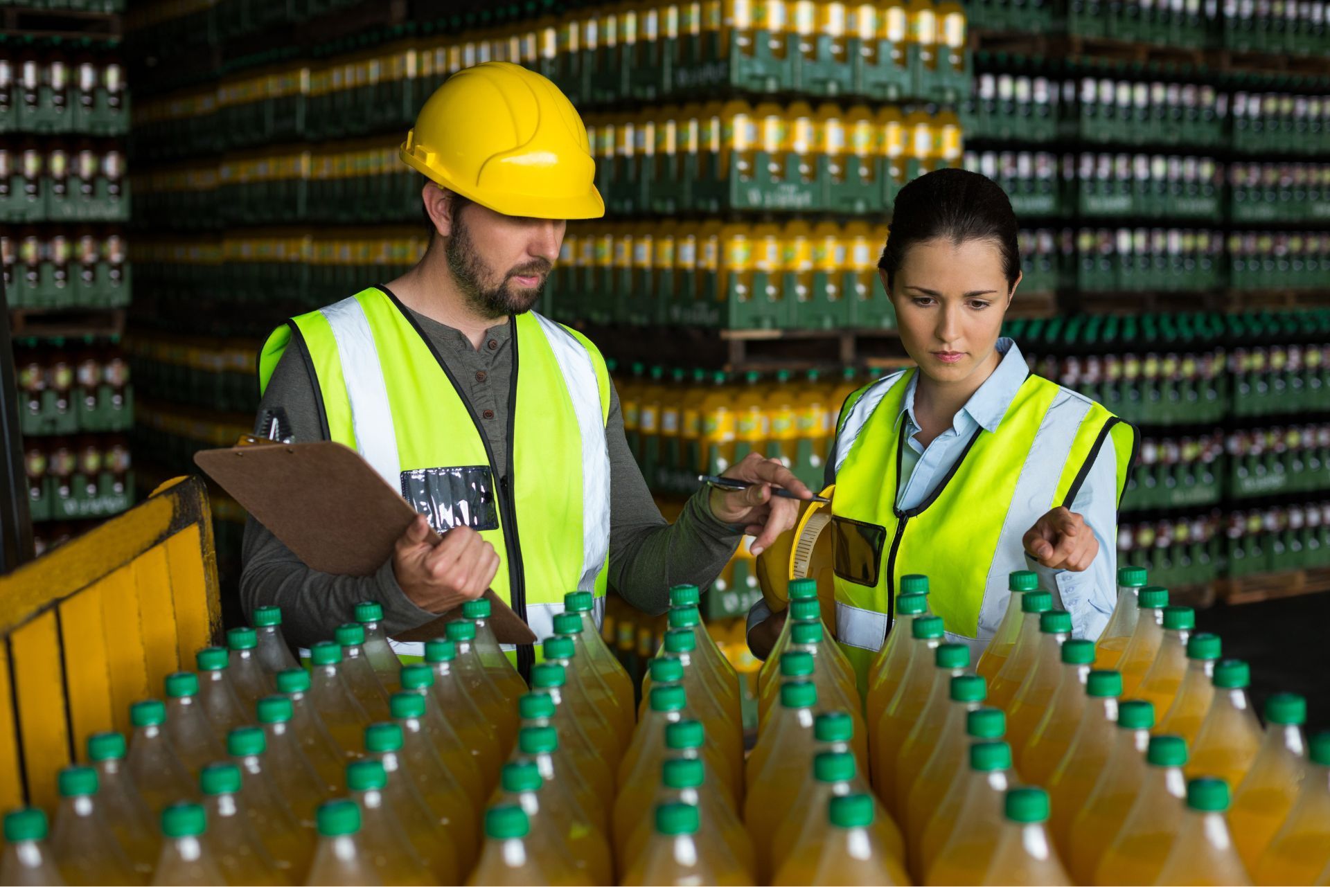 Two workers in safety vests and helmets inspecting rows of bottles in a warehouse.